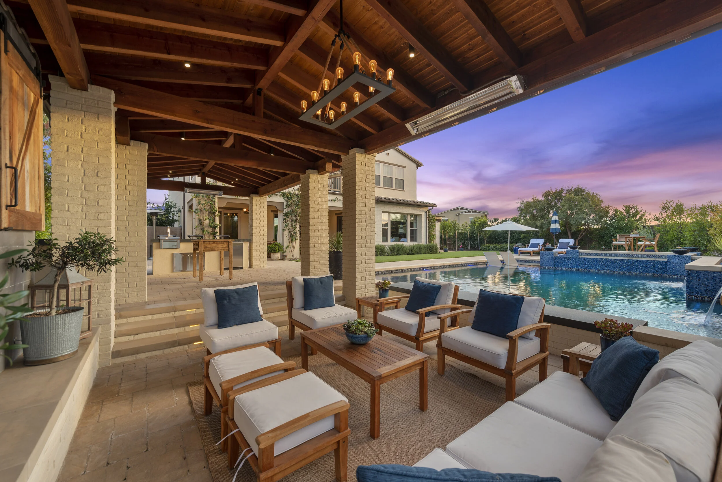 Covered patio with outdoor seating including white cushioned chairs and a sofa, a wooden coffee table with potted plants, overlooking a swimming pool with a waterfall feature and a sunset sky in the background.