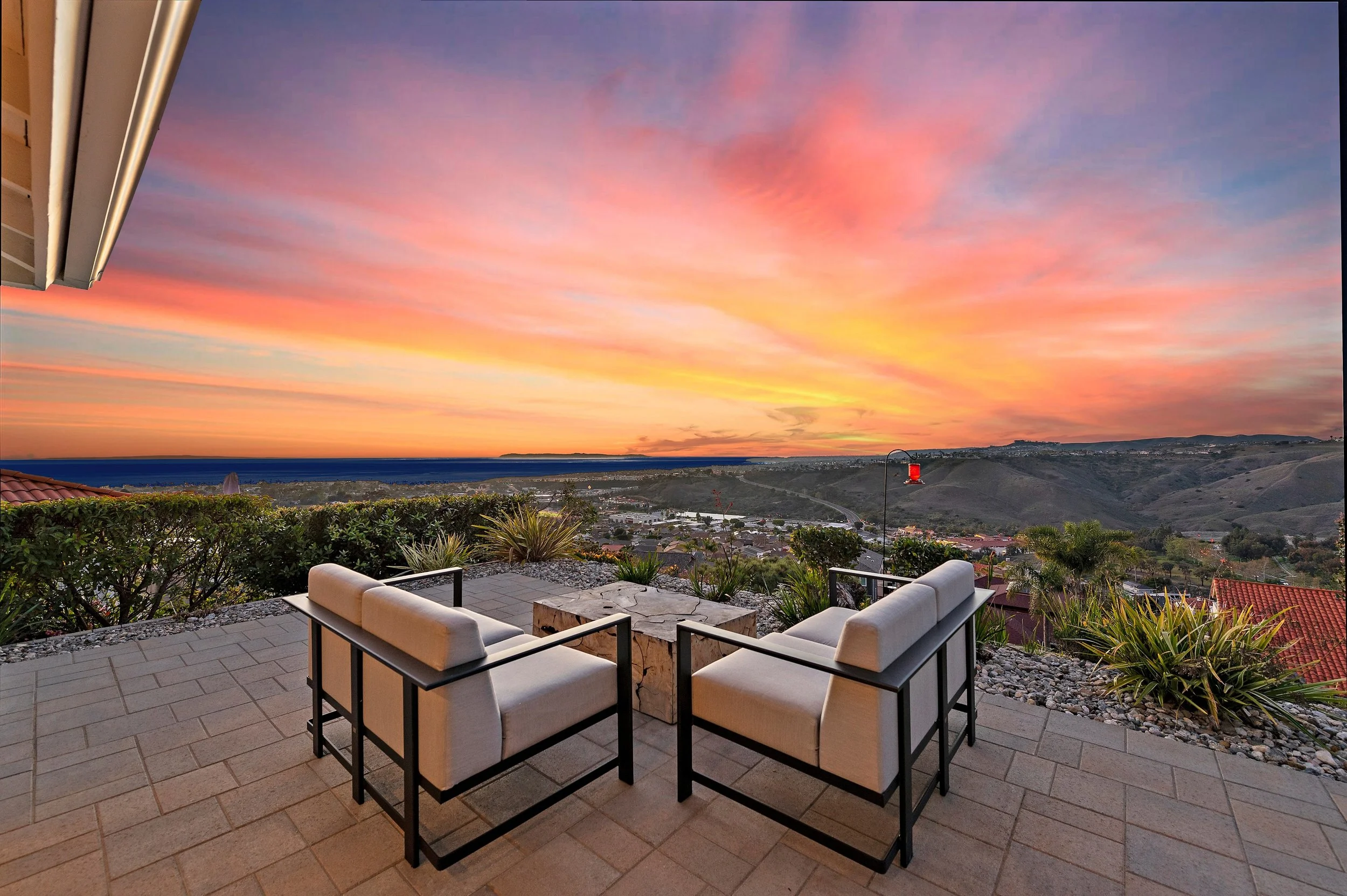 Patio with two white cushioned chairs facing a sunset over hills and a small town, with a view of the sky painted in pink, orange, and purple hues.