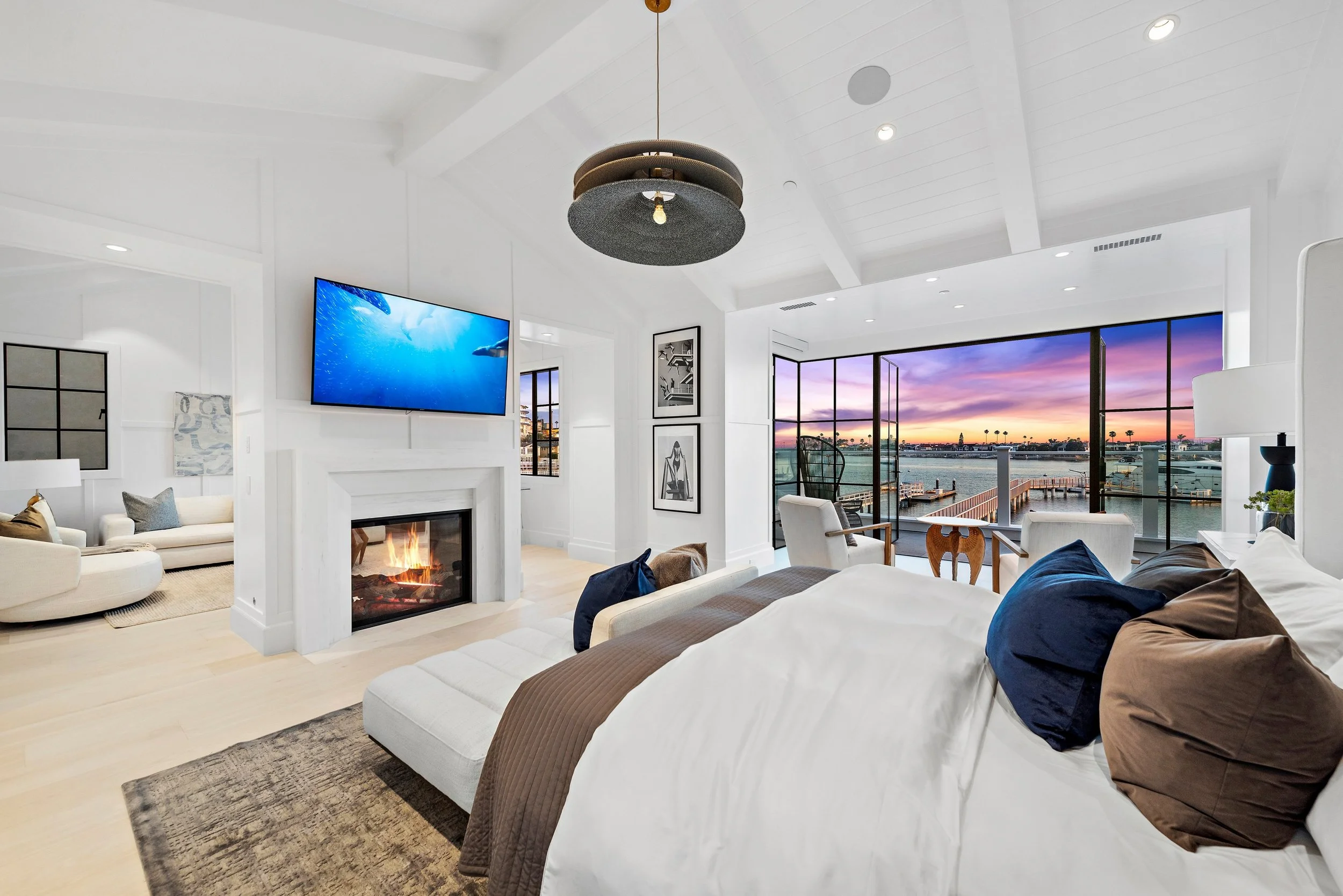 Modern bedroom with white walls, a fireplace, a mounted flat-screen TV, and large glass doors opening to a balcony with a view of the water and a sunset sky.