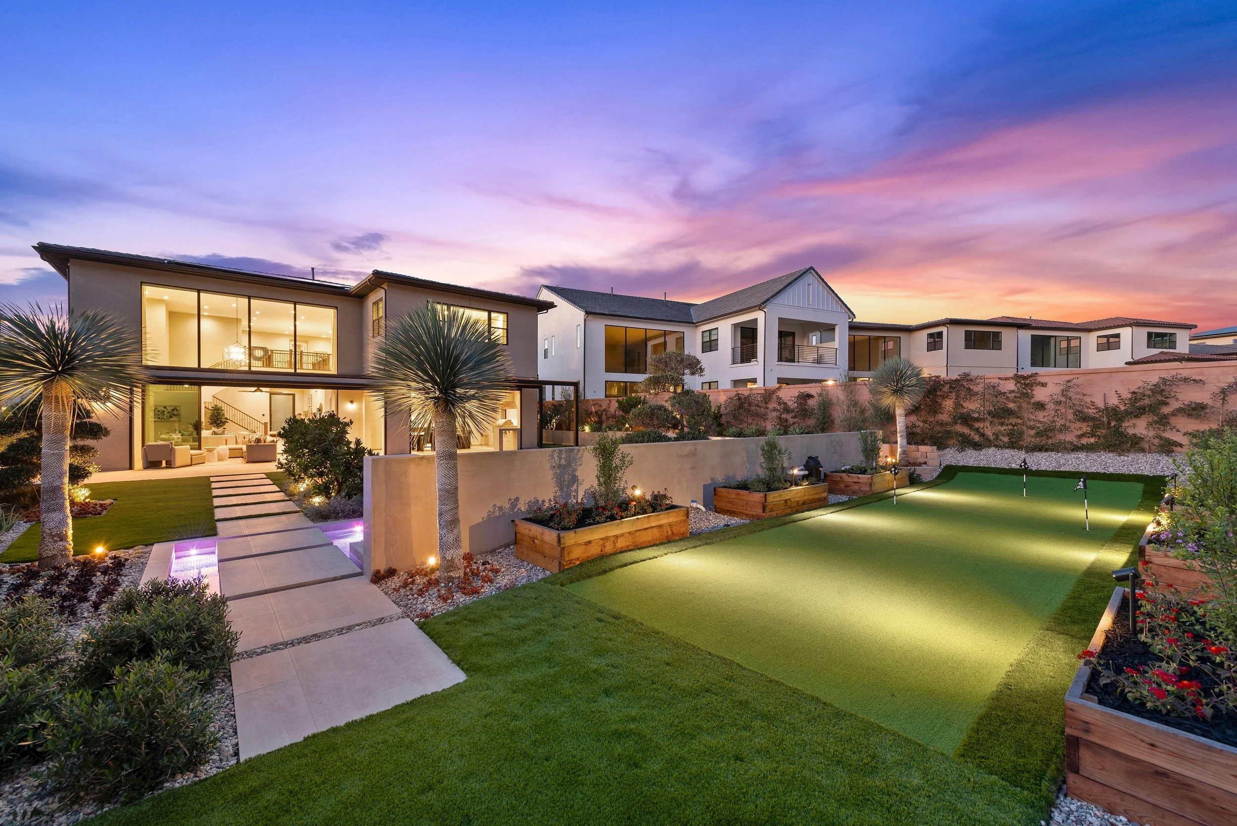 Modern backyard with a mini golf course, palm trees, landscaped plants, and a house with large glass windows at sunset.