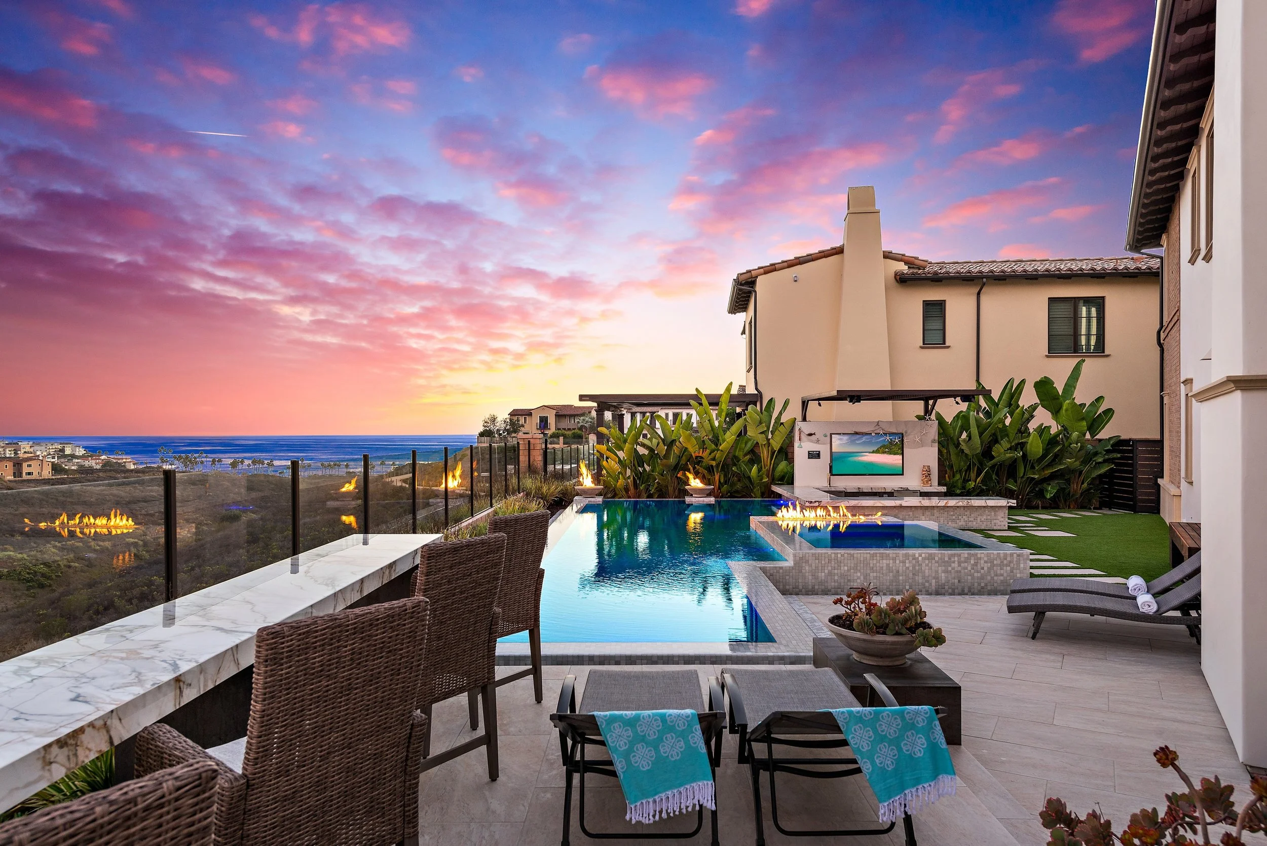 Luxury backyard with swimming pool, lounge chairs, outdoor TV, fire features, and ocean view at sunset.