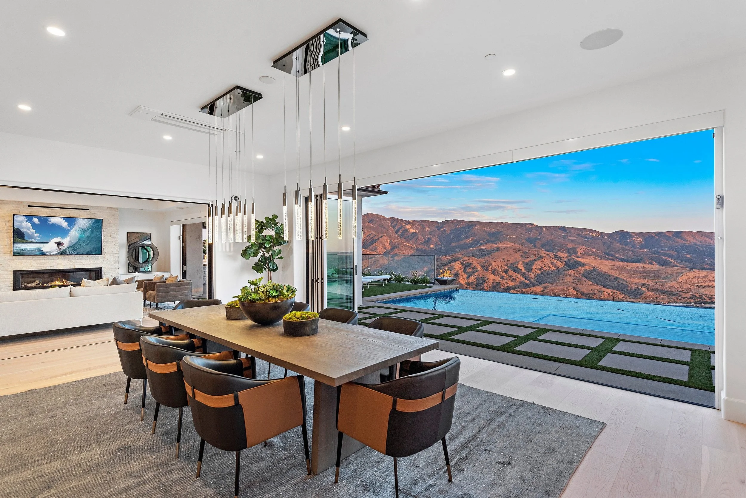 Modern open-concept living and dining area with a mountain view, large sliding glass door open to a pool and outdoor patio, stylish dining table with eight chairs, contemporary pendant lights, and a cozy living room with a fireplace.