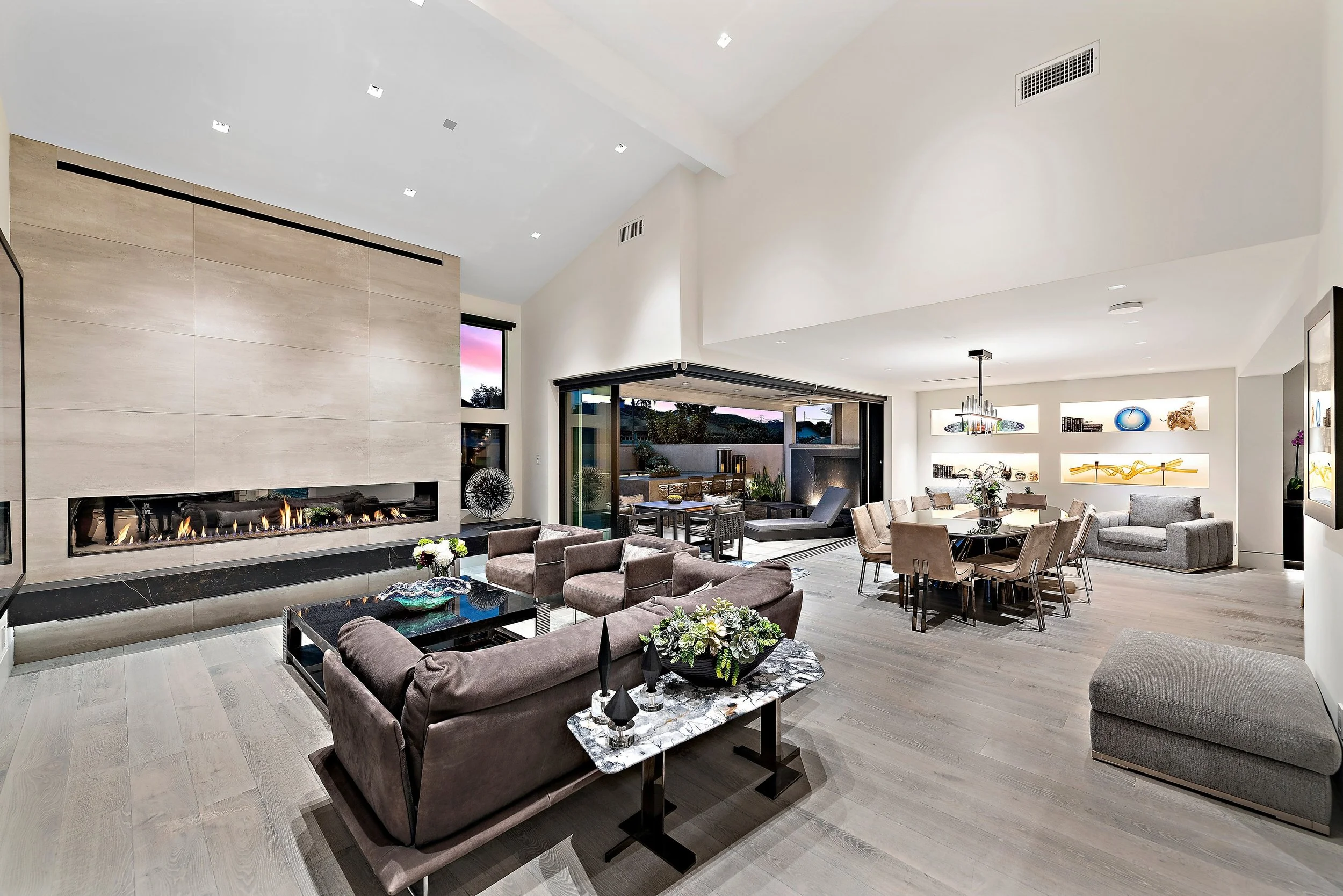 Spacious modern living room with a long gas fireplace, contemporary furniture, and an open kitchen with dining area, illuminated shelves, and large windows showing outdoor seating.