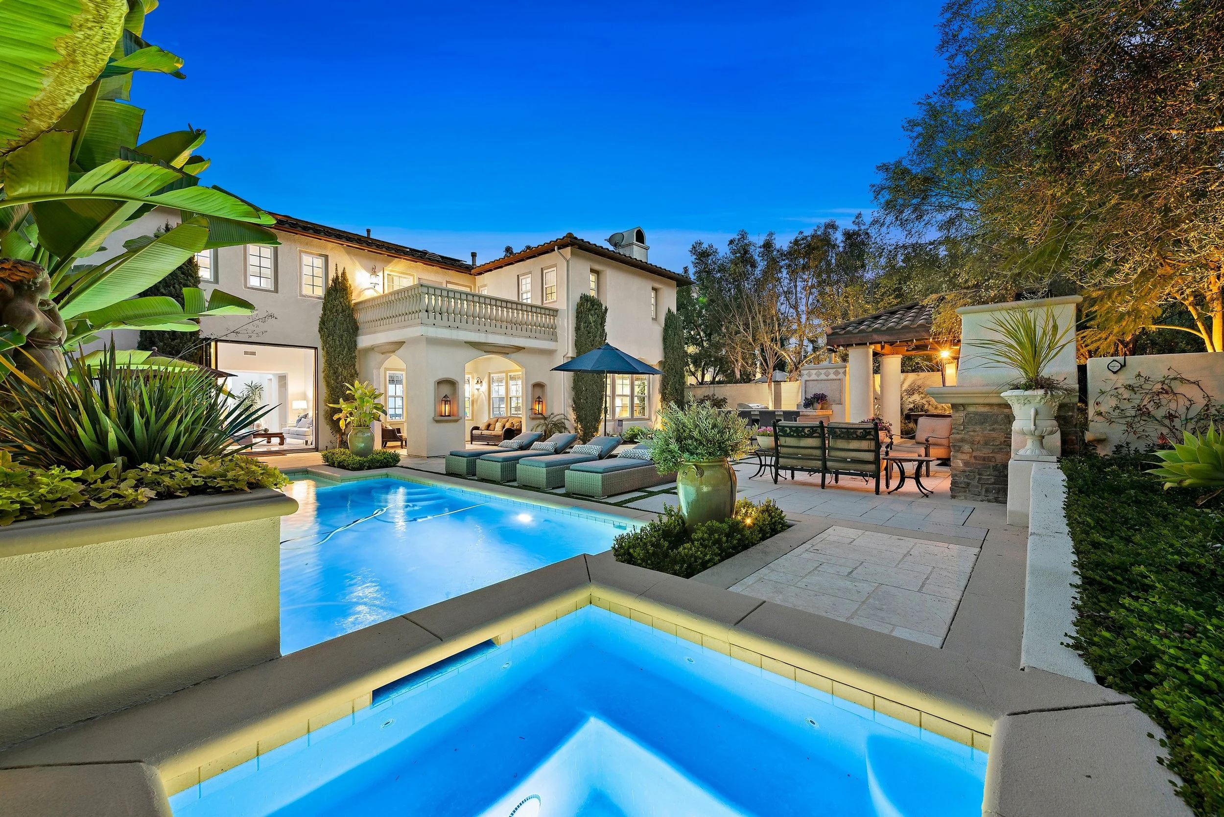Luxury backyard with illuminated pool, outdoor seating, and a white two-story house during twilight.