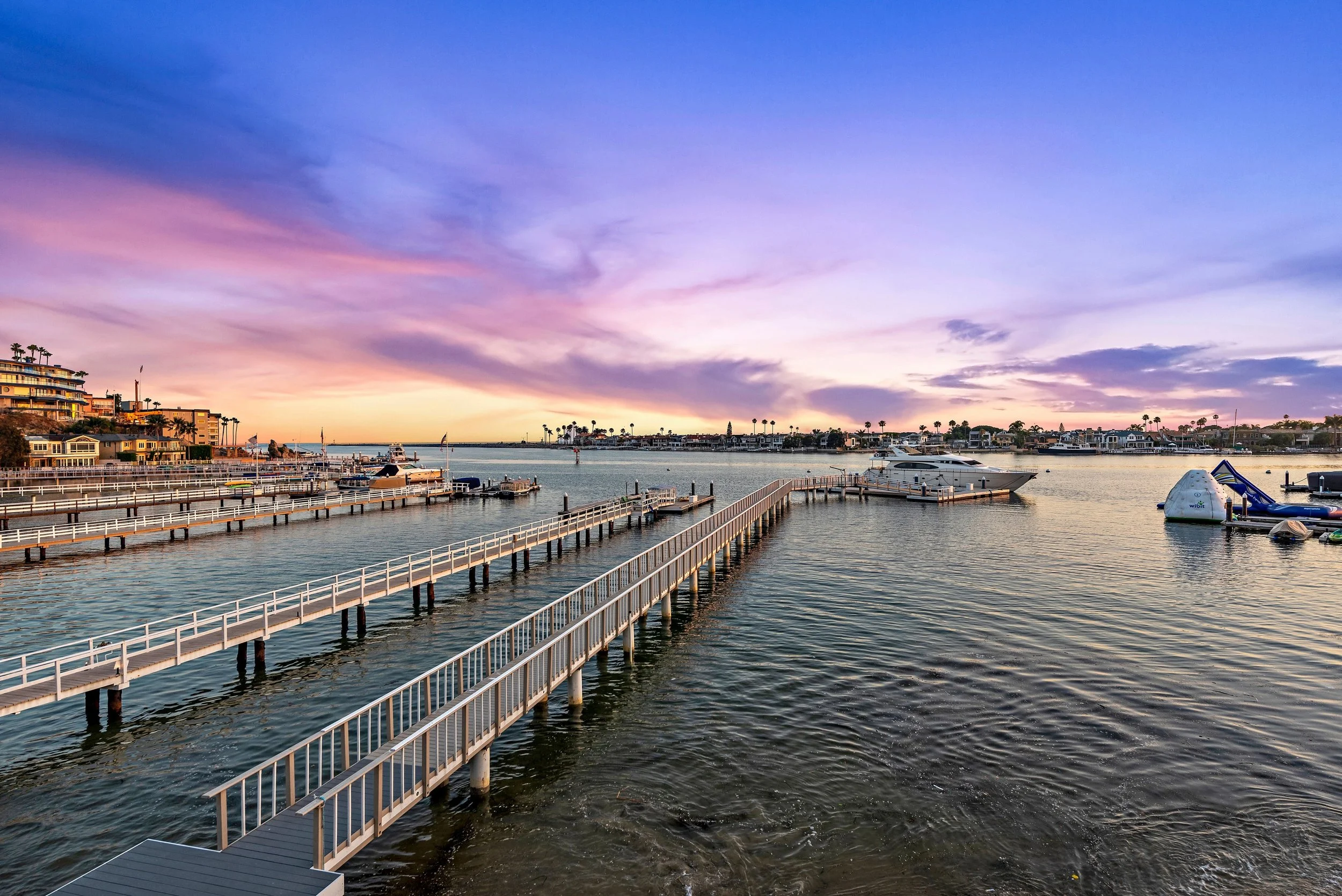Sunset over a marina with boats docked along piers, colorful sky with purple, pink, and orange hues, and houses along the shoreline.