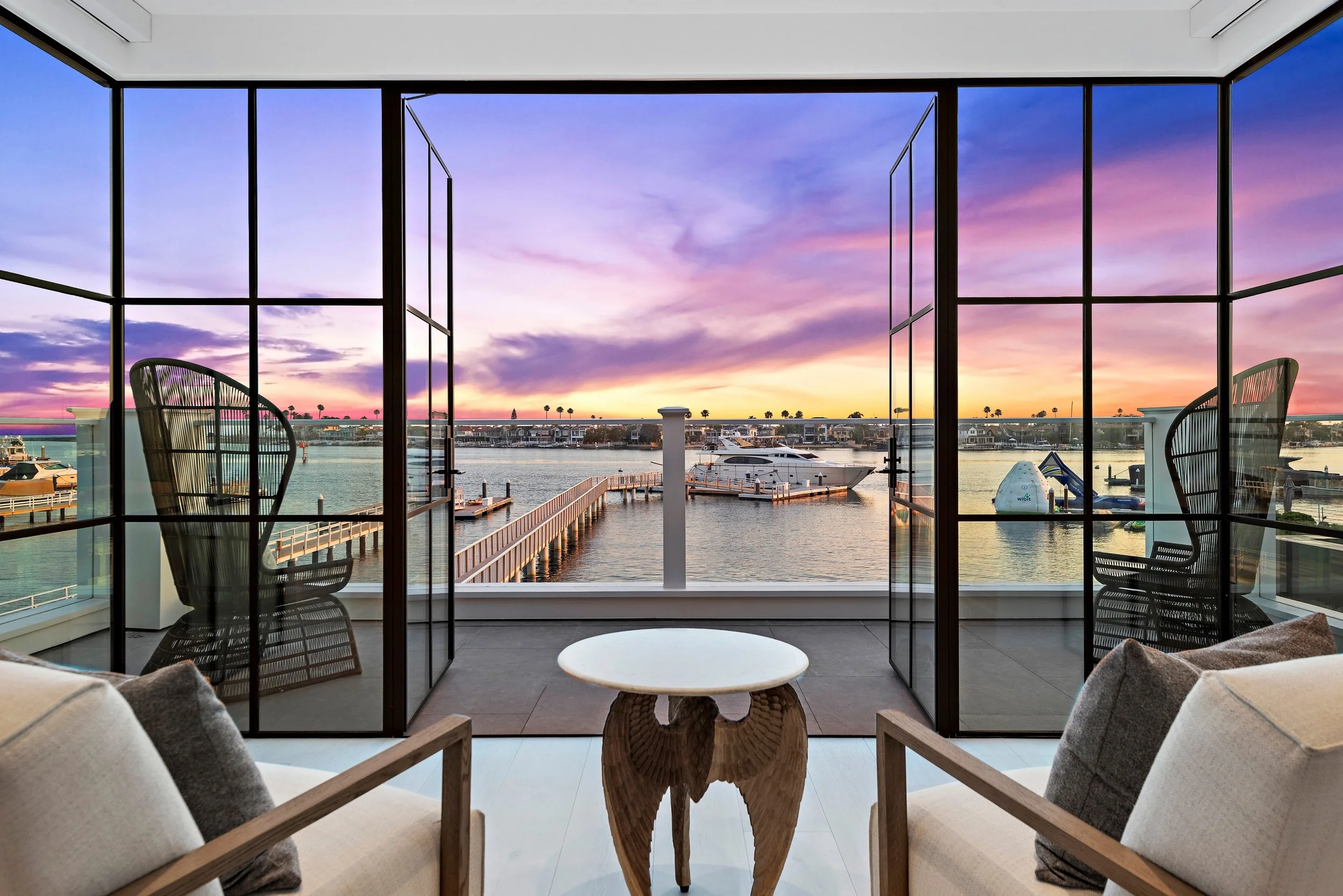 Living room with large open glass doors leading to a balcony overlooking a marina at sunset, with boats and water visible.