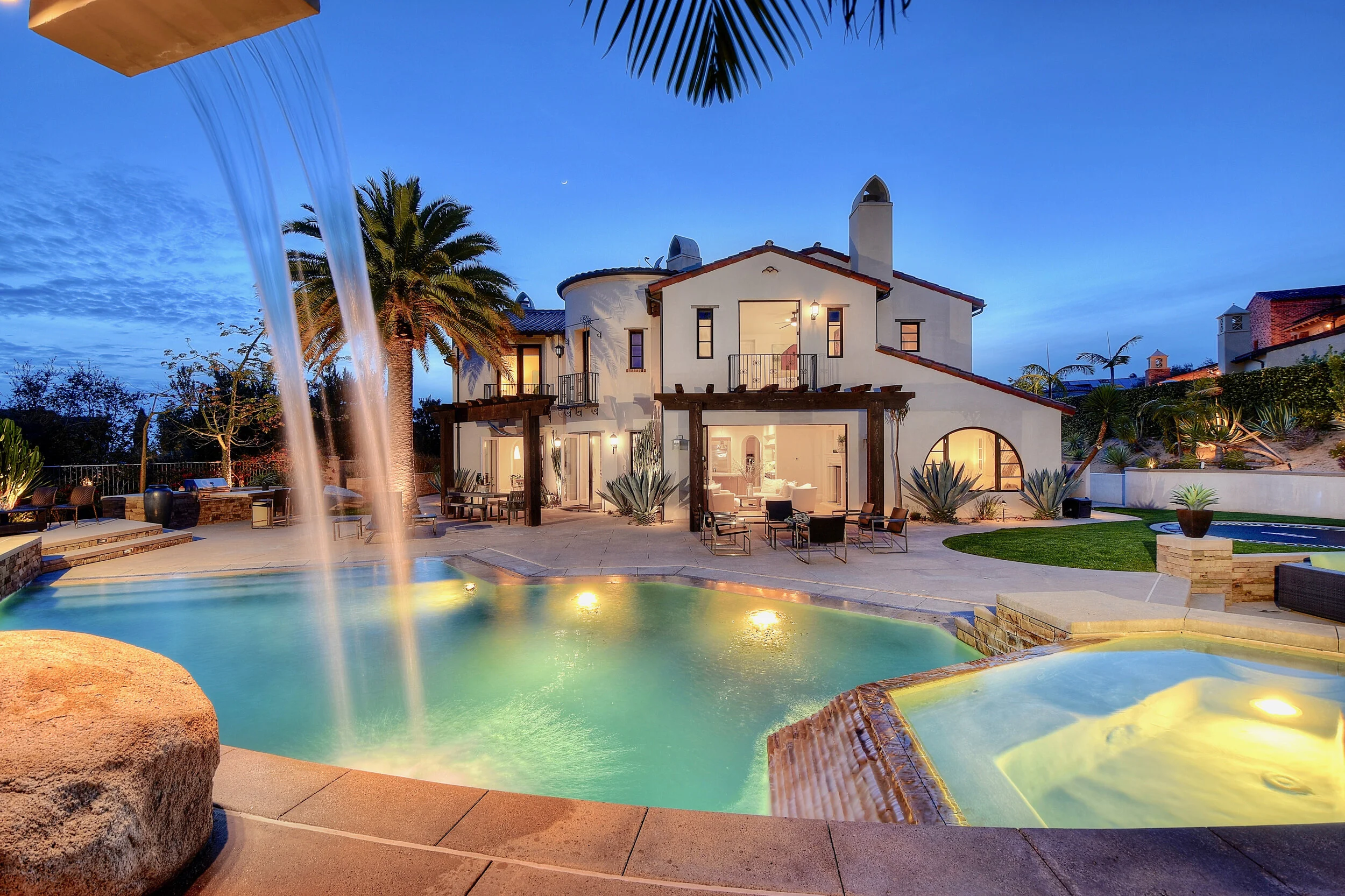 Luxury house with a swimming pool, waterfall feature, outdoor seating, and desert plants during dusk.