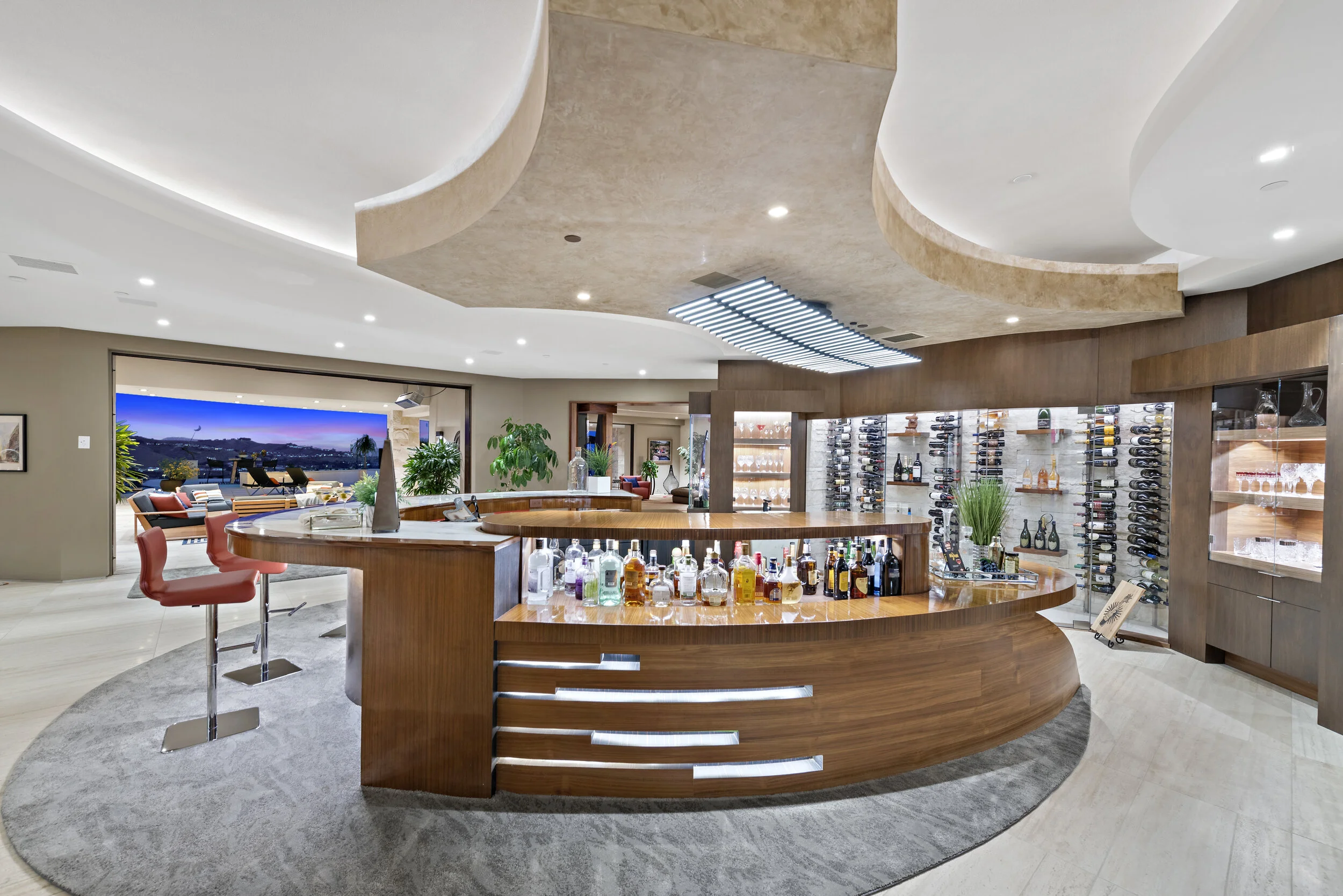 Modern home bar with a curved wooden counter, shelves of liquor bottles, wine racks, and a view of an outdoor patio with seating and a cityscape in the background.