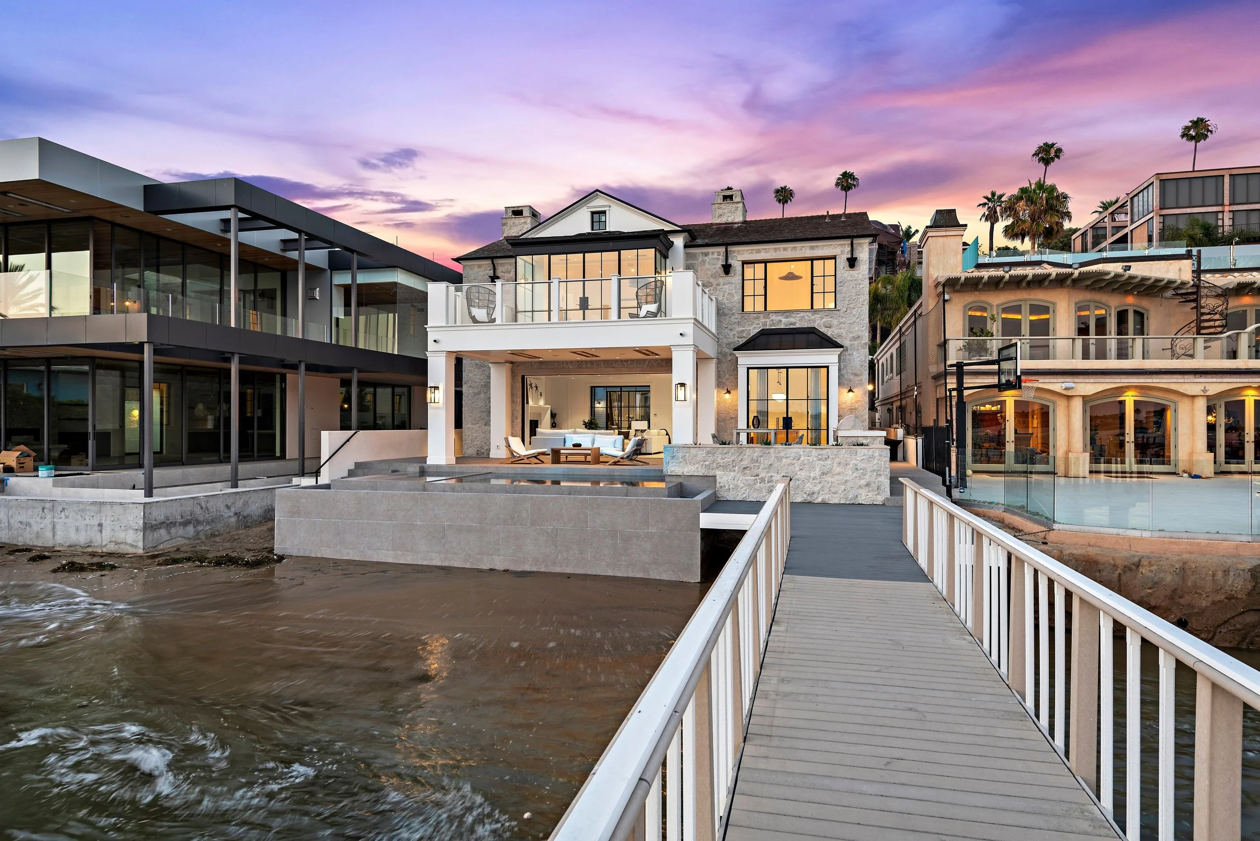Luxury beachfront houses during sunset with a dock leading to the water and a pool on the right.
