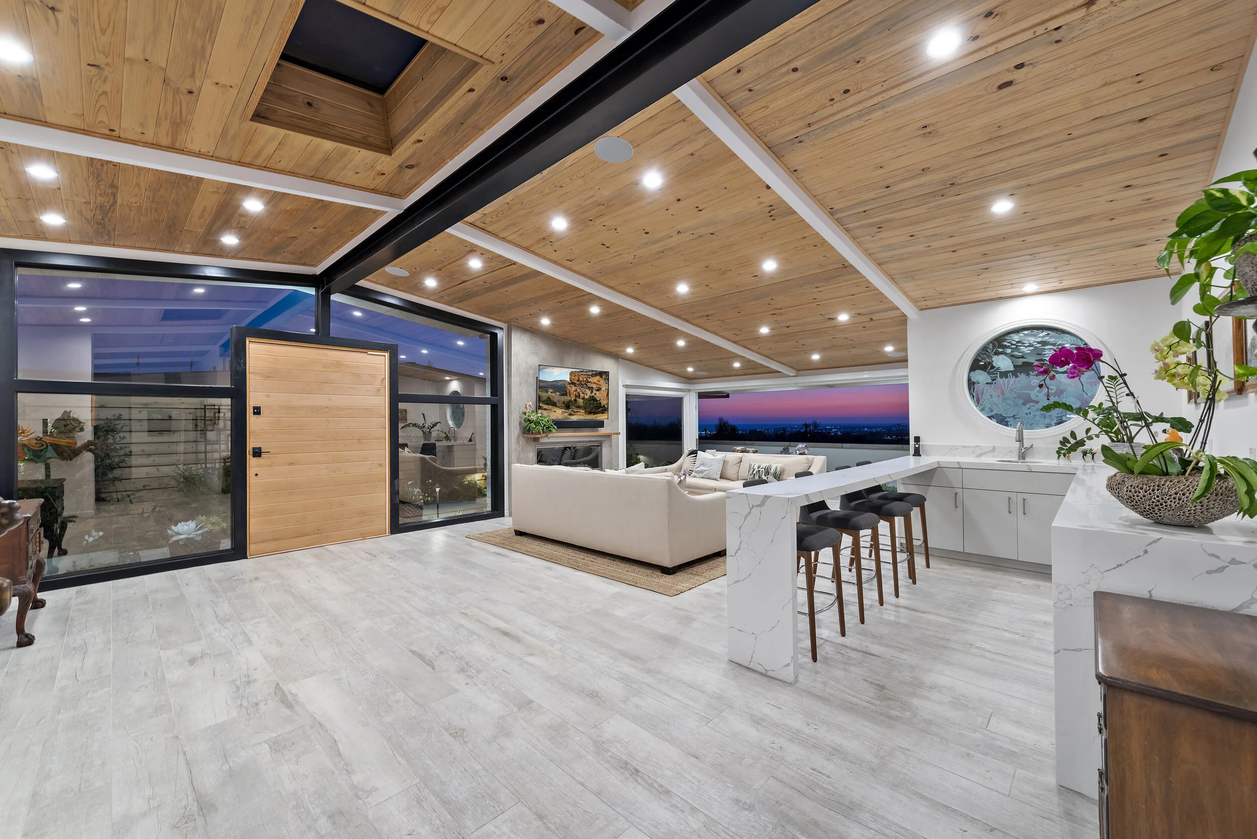 Modern open-concept living area with wooden ceiling, white marble kitchen island with barstools, beige sofa, and large windows showing a sunset view