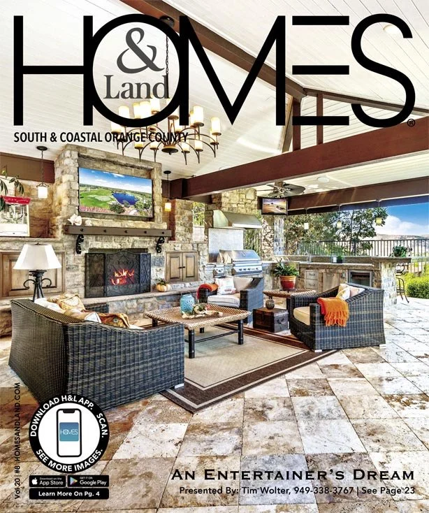 Cozy indoor living room with stone fireplace, flat-screen TVs, wicker chairs, a coffee table, outdoor view, and a chandelier, featured in H&O Homes magazine.