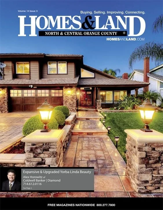 Front view of a two-story house with a brick and wood exterior, illuminated by pathway lamps, with a paved walkway and landscaped lawn, on the cover of Homes & Land magazine.