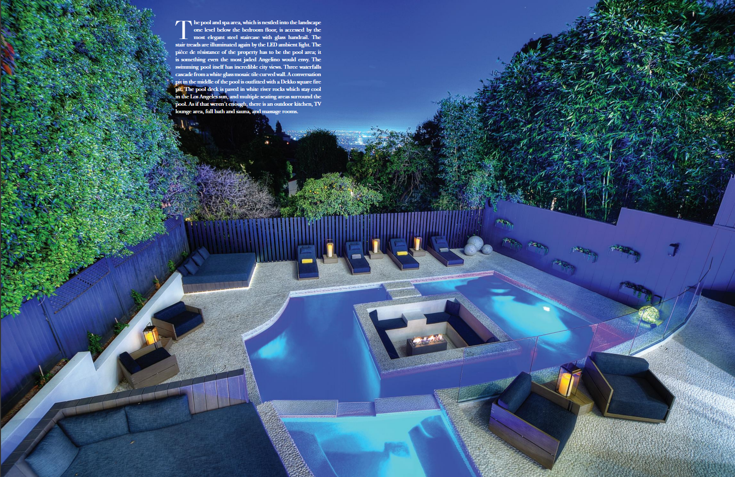 Nighttime view of a backyard pool area with multiple lounging chairs, a fire pit, and illuminated planters, surrounded by tall trees and a view of a distant city skyline.