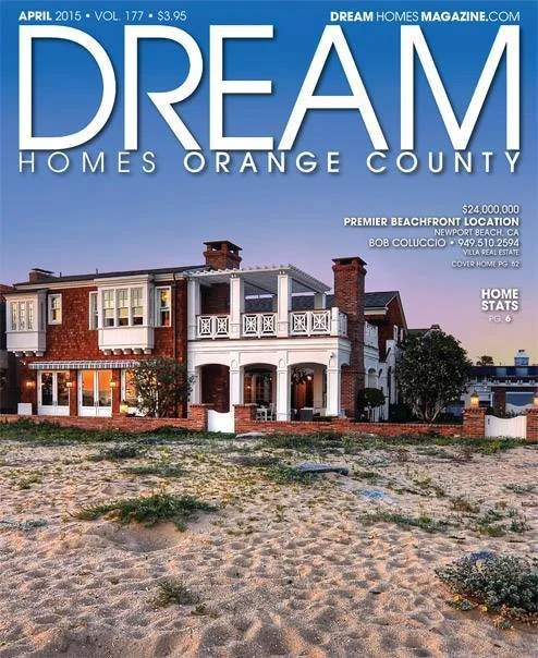 A large beachfront house with a brick and white exterior, multiple balconies, and a sandy area with sparse vegetation in the foreground. The sky is clear with a gradient from pinkish to blue.