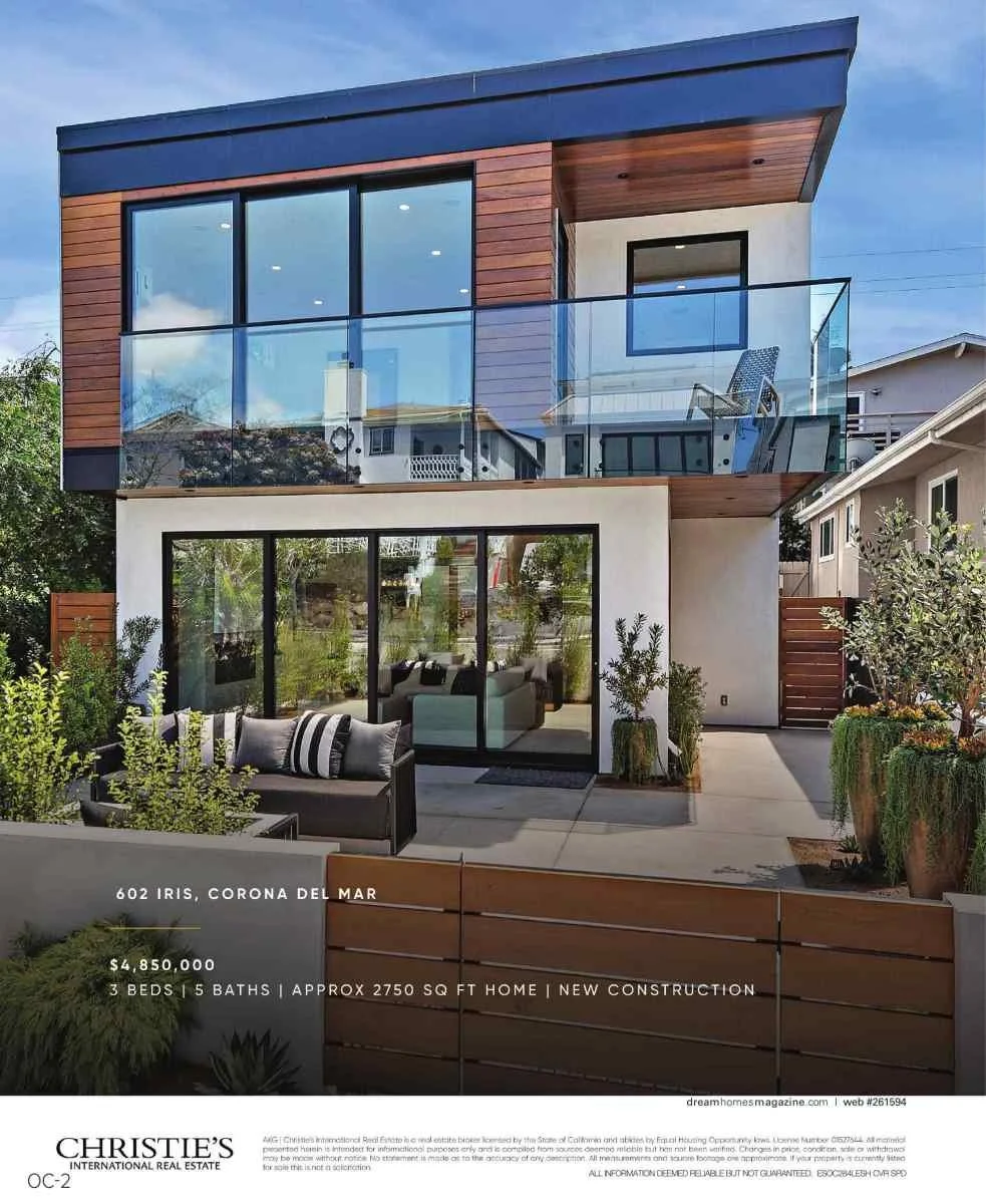 Modern three-story house with large glass windows, a balcony on the second floor, and a patio with outdoor furniture, surrounded by plants.