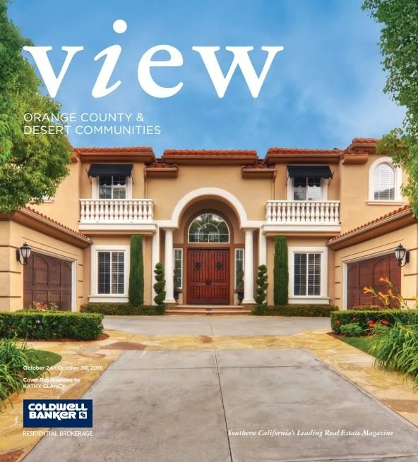 Cover of a real estate magazine featuring a large, luxurious two-story house with an arched entrance, front porch, and two garages, set against a blue sky with trees on each side.