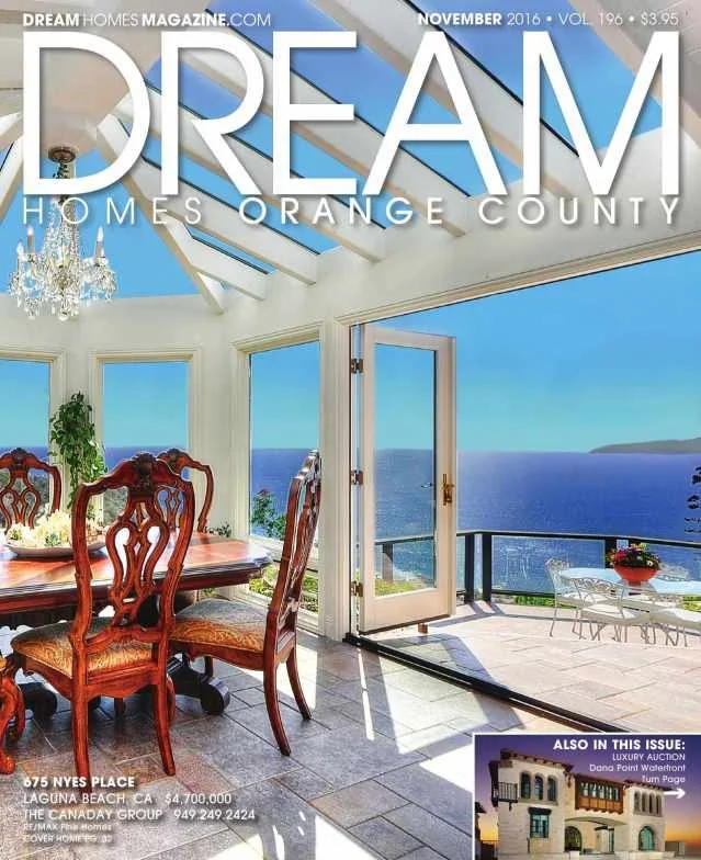 A bright dining area with large glass windows and door leading to a balcony overlooking the ocean. Wooden dining table with ornate chairs, white walls, chandelier, and skylights in a house built in a modern coastal style.