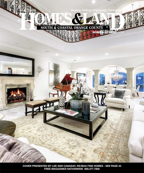 Luxurious living room with a fireplace, grand piano, large flat-screen TV, plush white sofas, dark wood coffee table with books and decorative items, large window with views, and elegant chandelier.