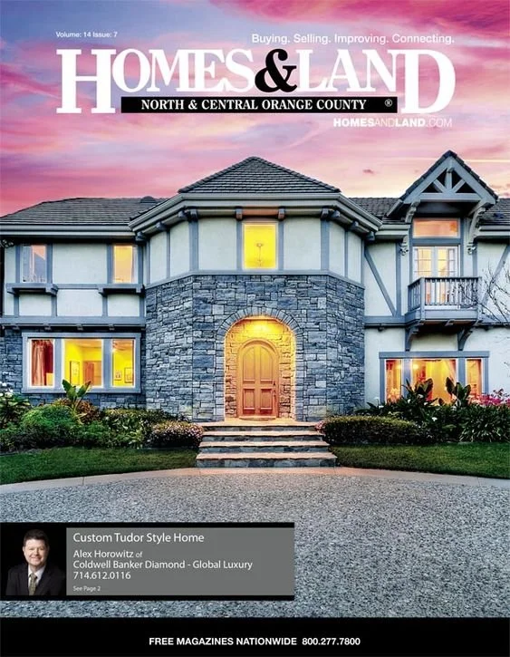 Cover of Homes & Land magazine featuring a large, elegant Tudor-style house with stone and stucco exterior, illuminated front door, well-maintained landscaping, and a colorful sunset sky in the background.