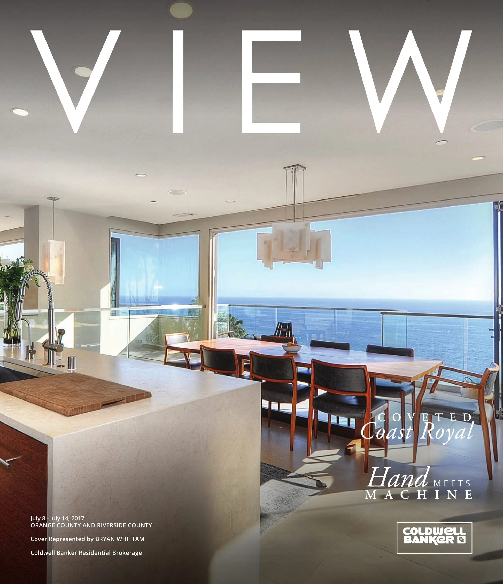 Modern dining area with a large wooden table, black and wooden chairs, and a view of the ocean through large windows. There is a white kitchen island with a cutting board and a sink on the left. The room has a minimalist design with pendant lighting 