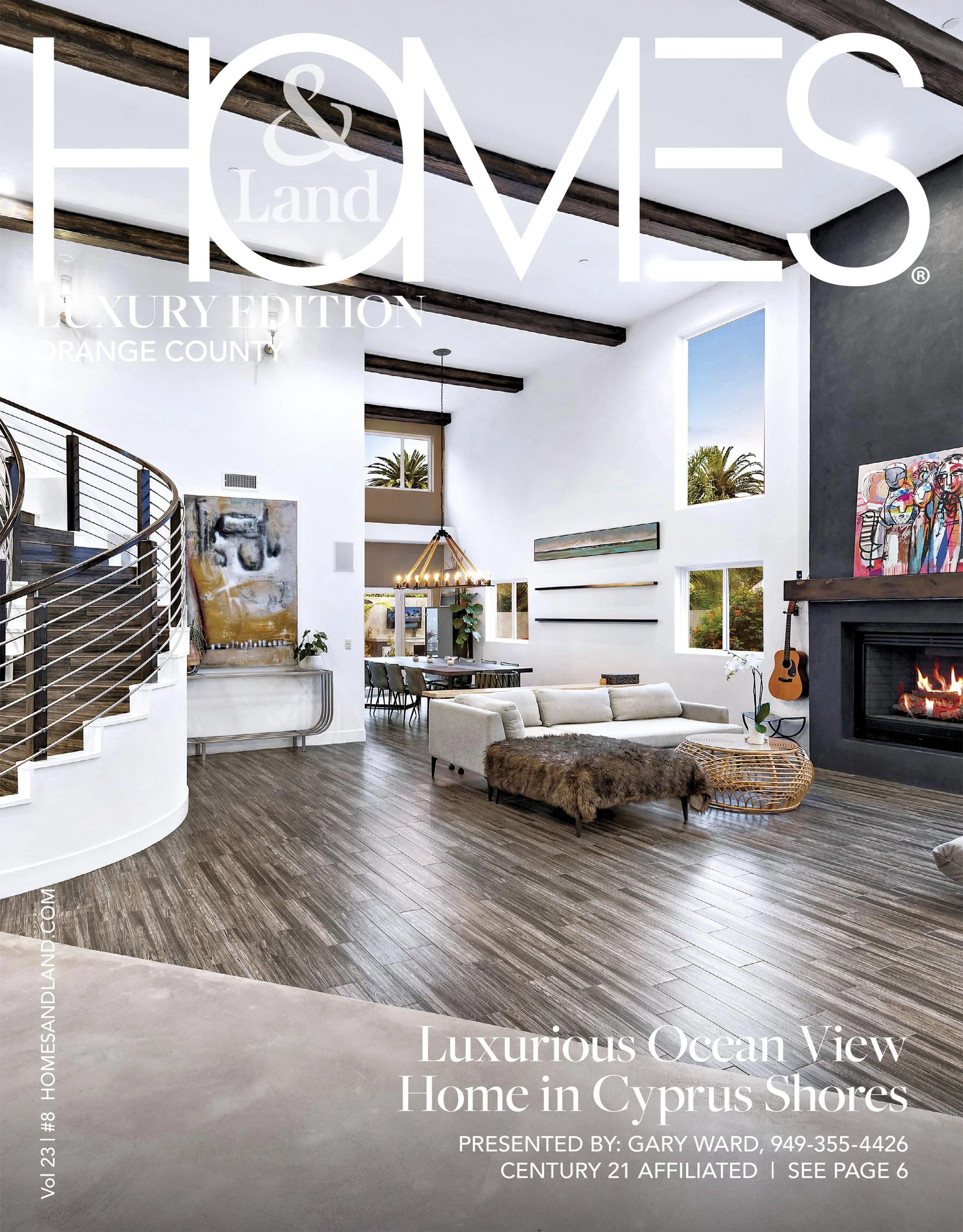 Cover of Homes & Land magazine featuring a spacious modern living room with high ceilings, wooden beams, large windows showing ocean views, white walls, contemporary furniture, a fireplace, and a staircase on the left side.