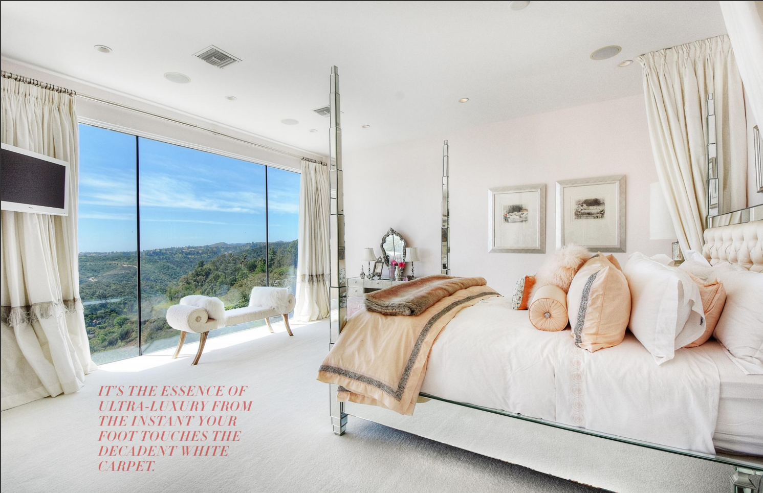 Luxurious bedroom with a glass wall view of green hills, white carpet, elegant bed with white and peach bedding, and a small bench outside the window.