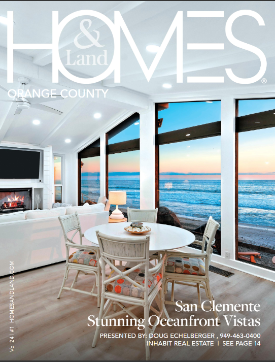 A bright, modern oceanfront living room with large windows showing a sunset over the ocean, a white sofa, a round dining table with four chairs, a fireplace, and a ceiling fan.