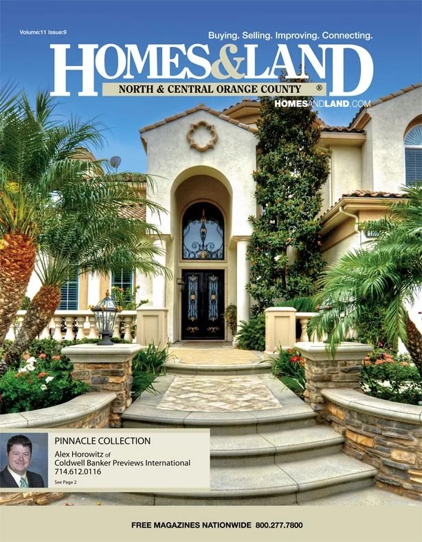 Front entrance of a luxury house with stone steps, lush tropical plants, and a wrought iron front door. The house has a beige stucco exterior and a tiled roof.