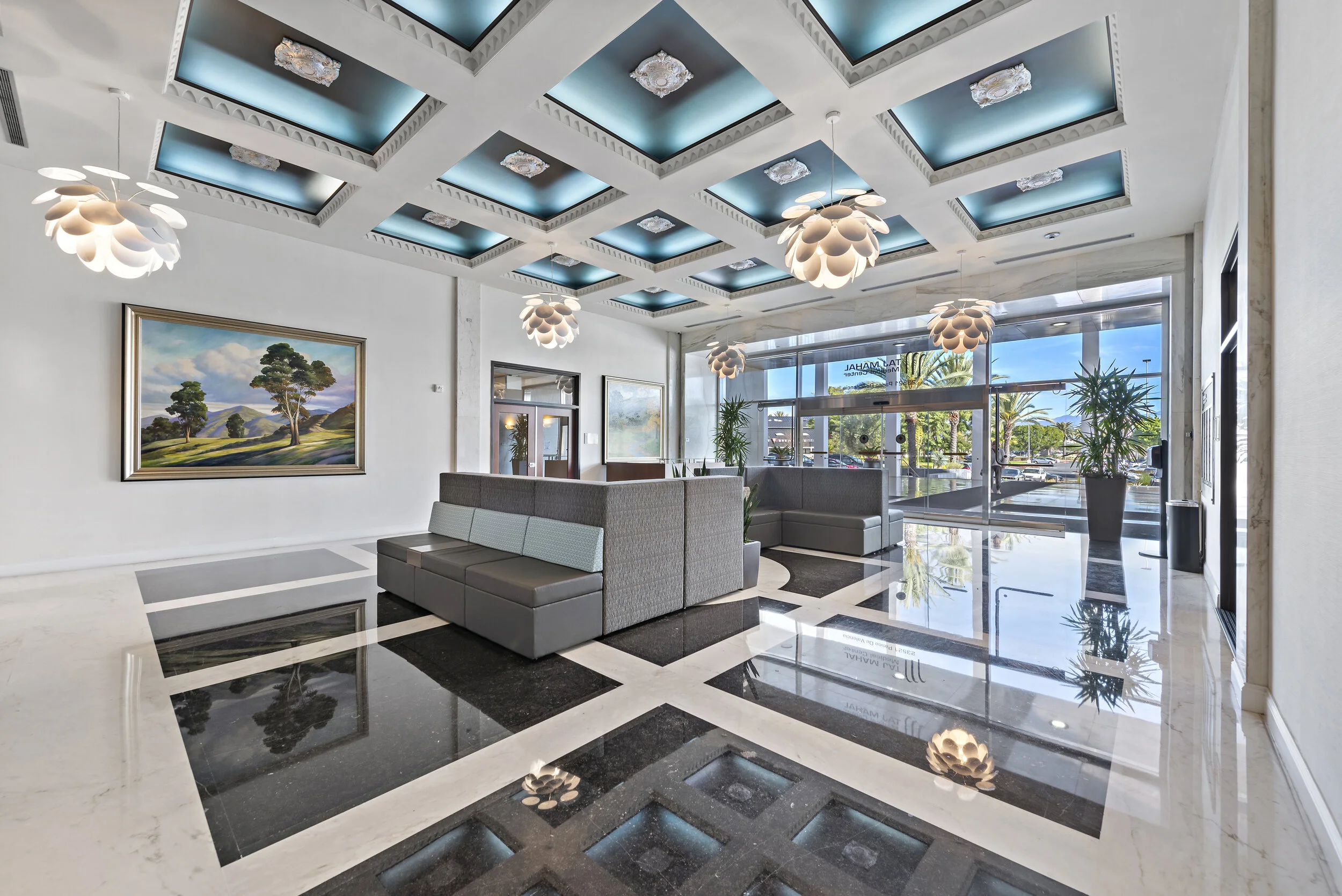 Modern hotel lobby with high ceiling, decorative lighting fixtures, large windows with outdoor view, potted plants, and sleek seating area with gray benches and black marble floor accents.