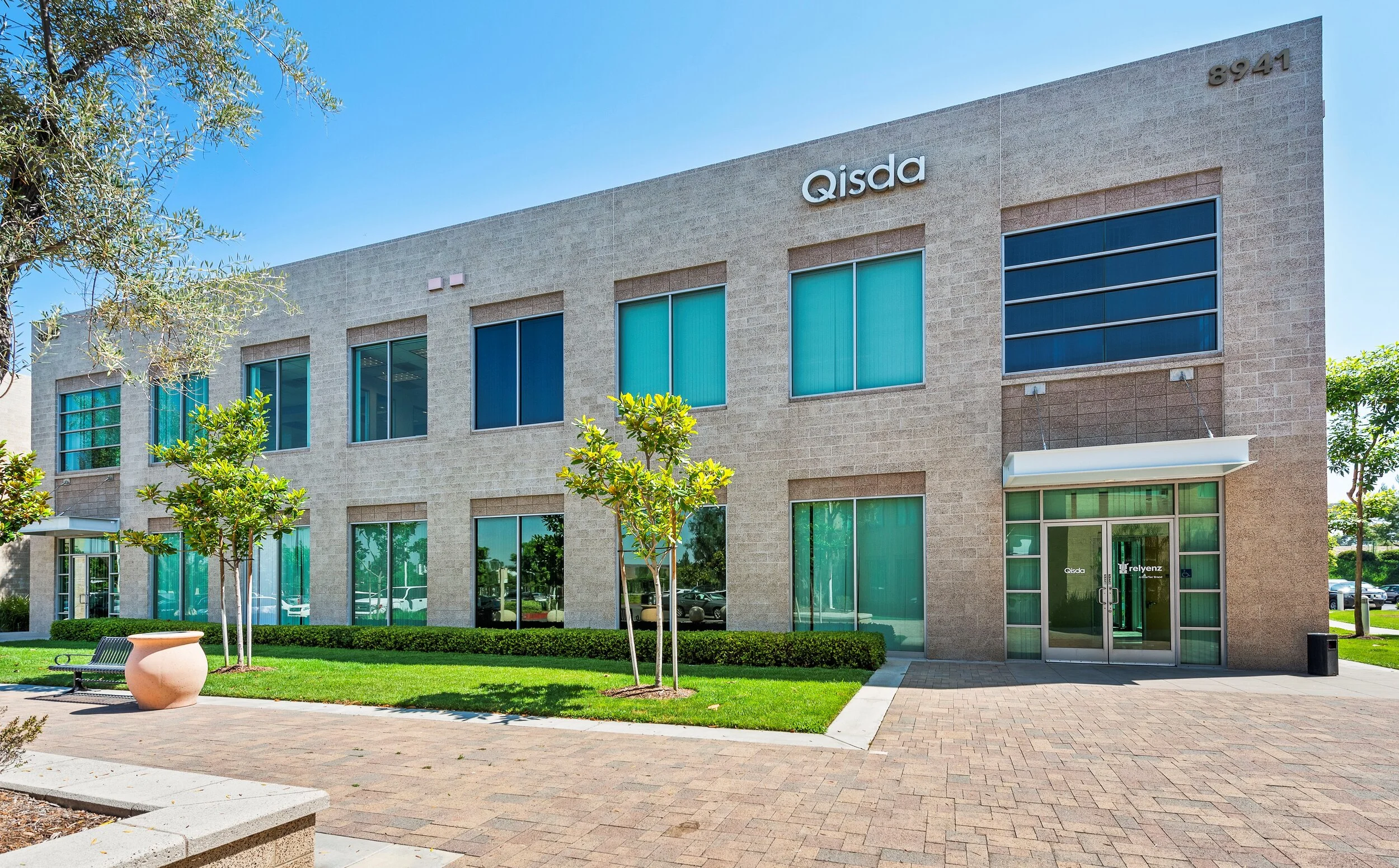 Exterior of a modern office building with the sign 'Qisda' above the entrance, surrounded by green trees, bushes, a sidewalk, and a blue sky.