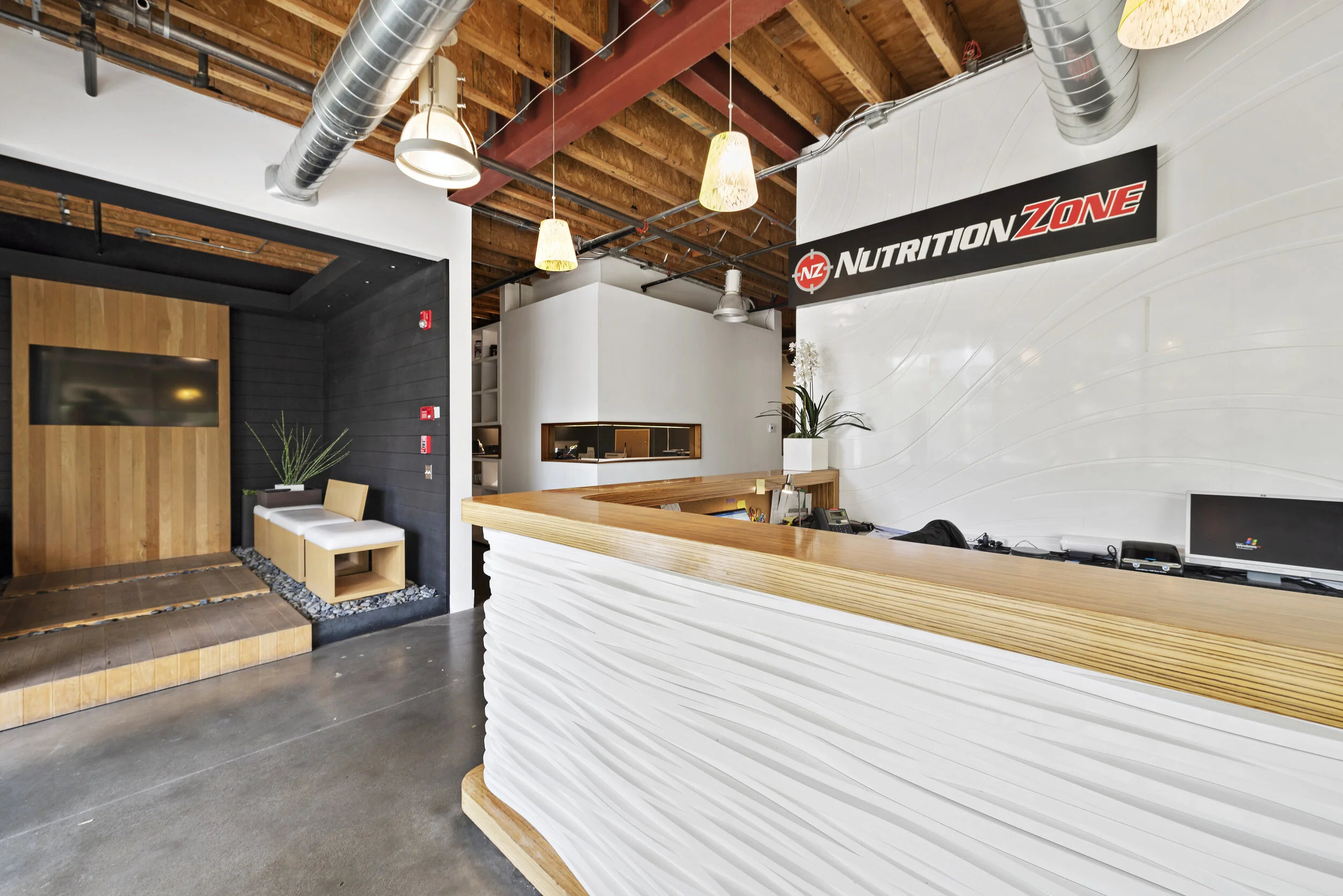 Modern reception area with a white textured reception desk, wooden accents, black and white walls, hanging pendant lights, and exposed ceiling ductwork.