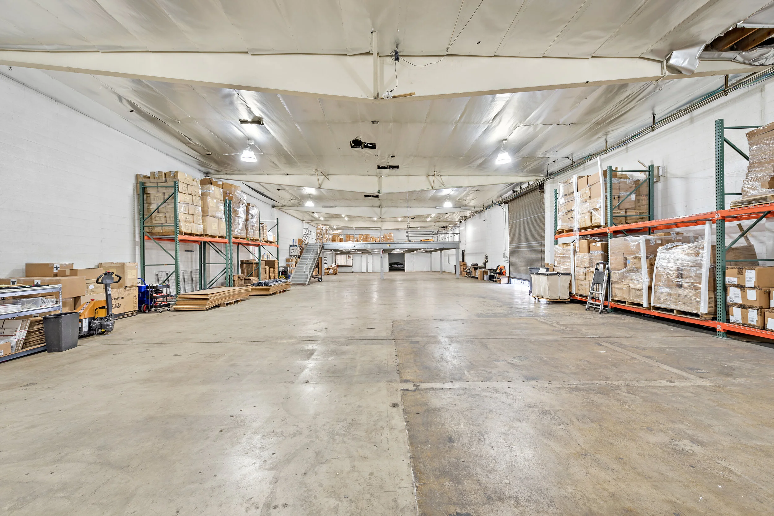 Empty warehouse with tall shelves stocked with cardboard boxes, a sealed roller door, and a mezzanine level in the background.