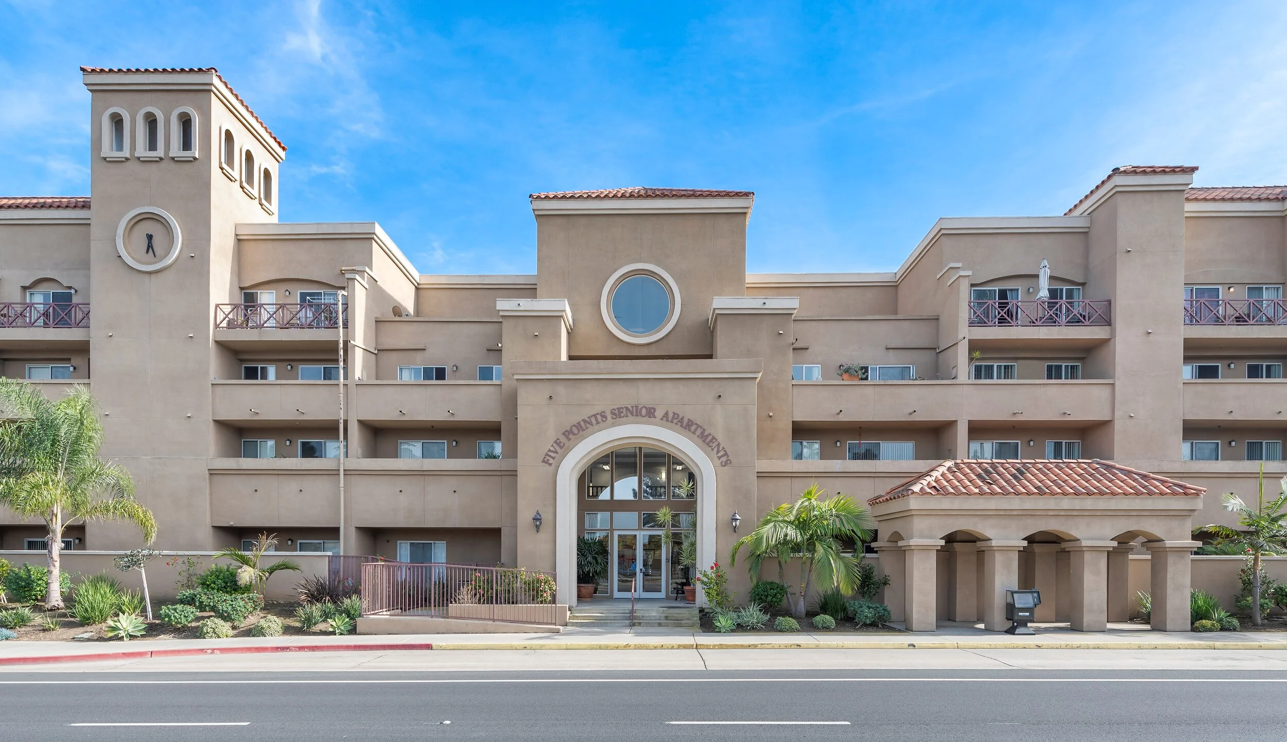 Front view of a beige senior apartment building with a sign that reads 'Five Points Senior Apartments', featuring balconies, a clock tower, and landscaped plants, under a blue sky.