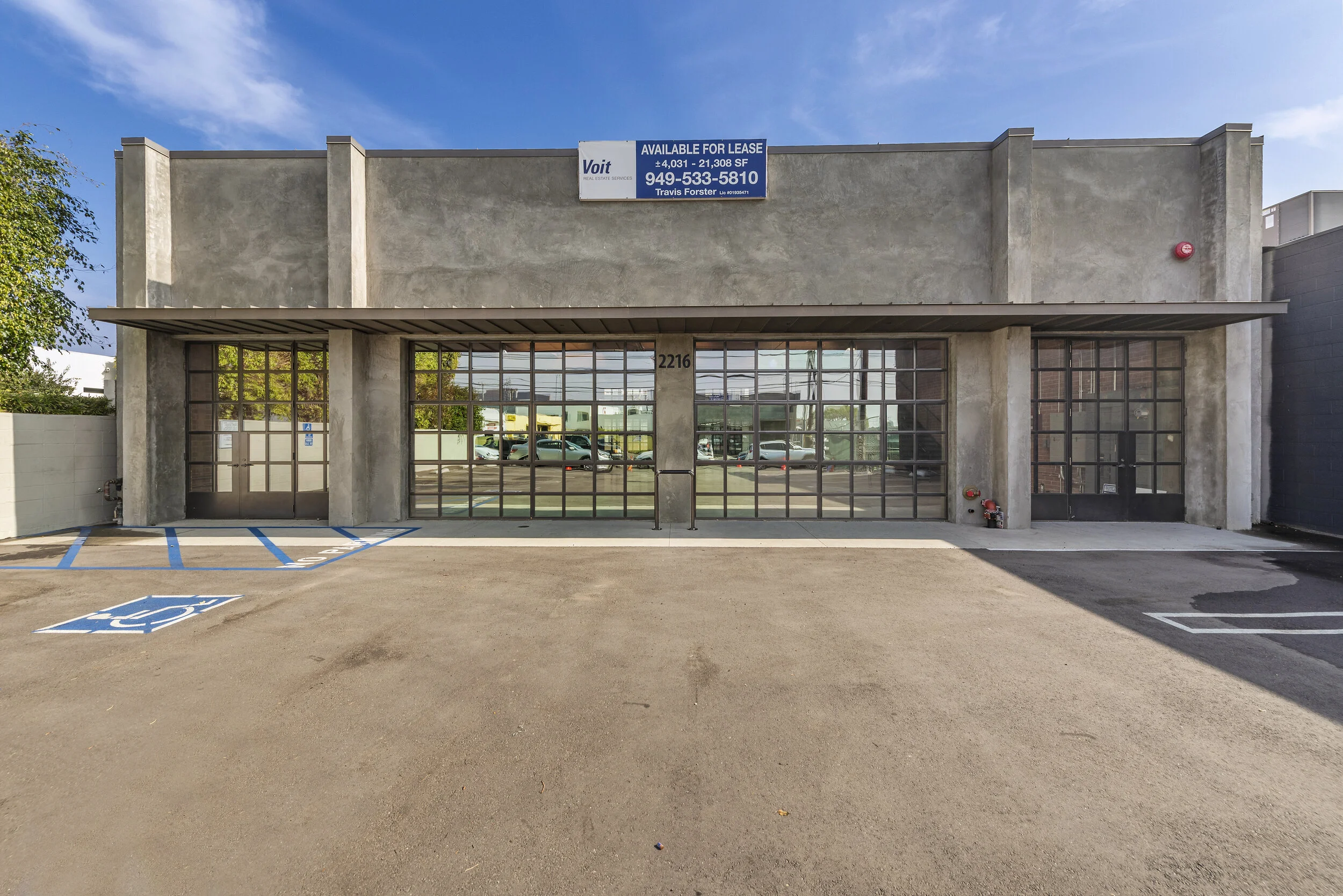 Commercial building with large glass windows and an available for lease sign, parking lot with designated Handicap accessible parking spot, and a clear blue sky.