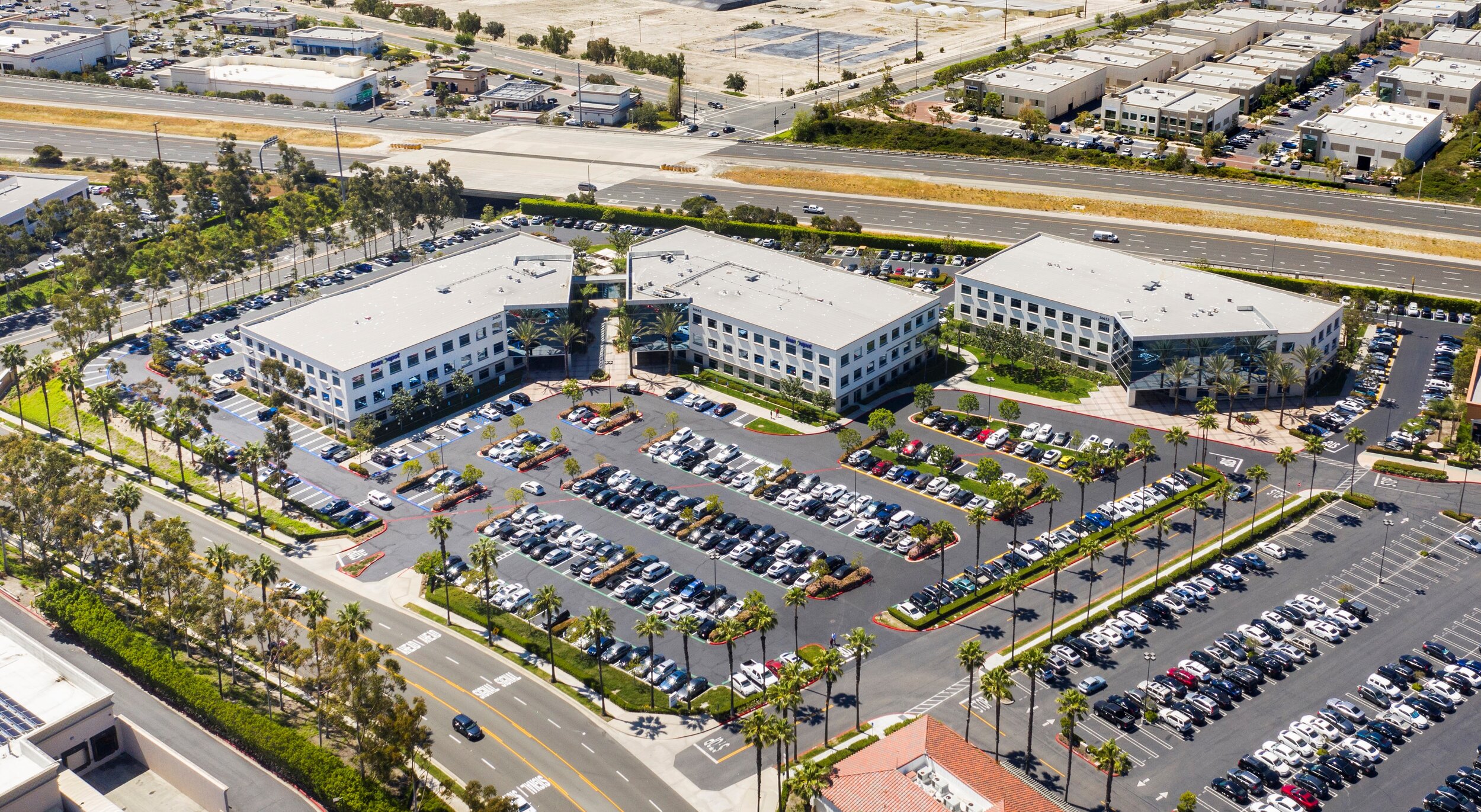 Aerial view of a commercial office complex with three white buildings, surrounded by parking lots filled with cars, palm trees, and nearby roads.