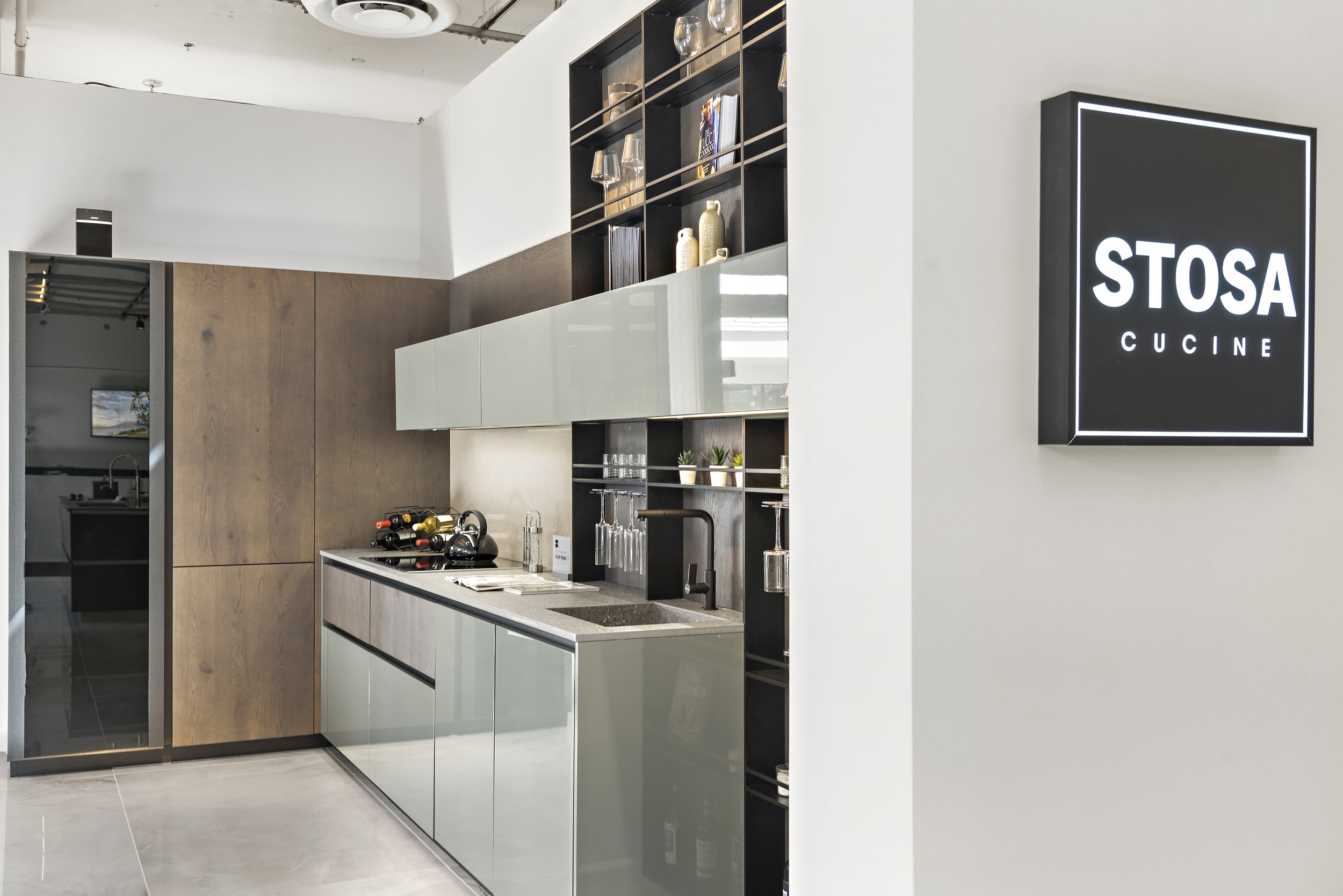 Modern kitchenette with light gray cabinets, black open shelves with glassware and books, a wooden wall panel, and a black illuminated sign reading 'STOSA CUCINE' on a white wall.