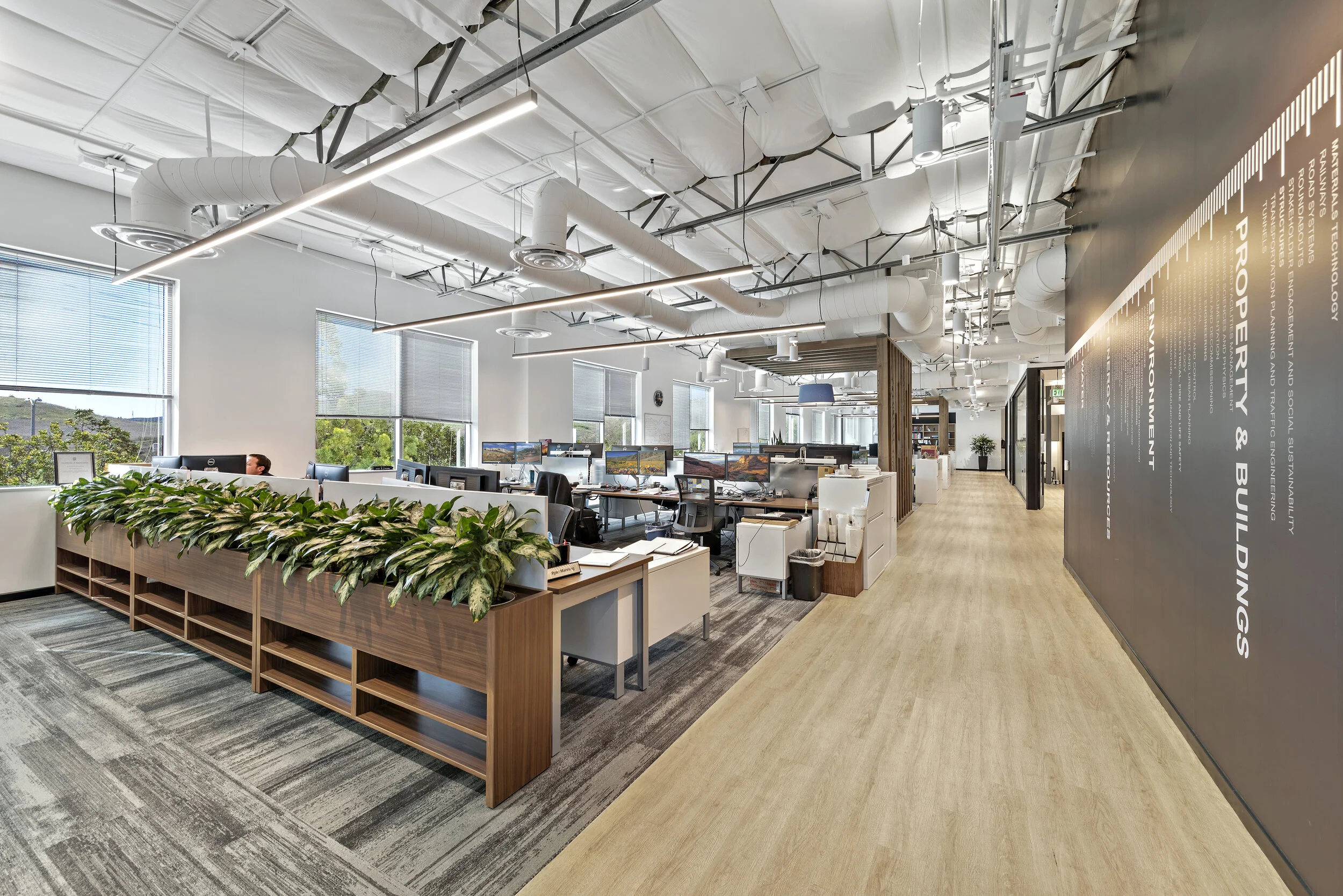 Modern open-plan office with multiple workstations, large windows with blinds, ceiling ducts, and a black wall with white text related to sustainability and environmental building topics.