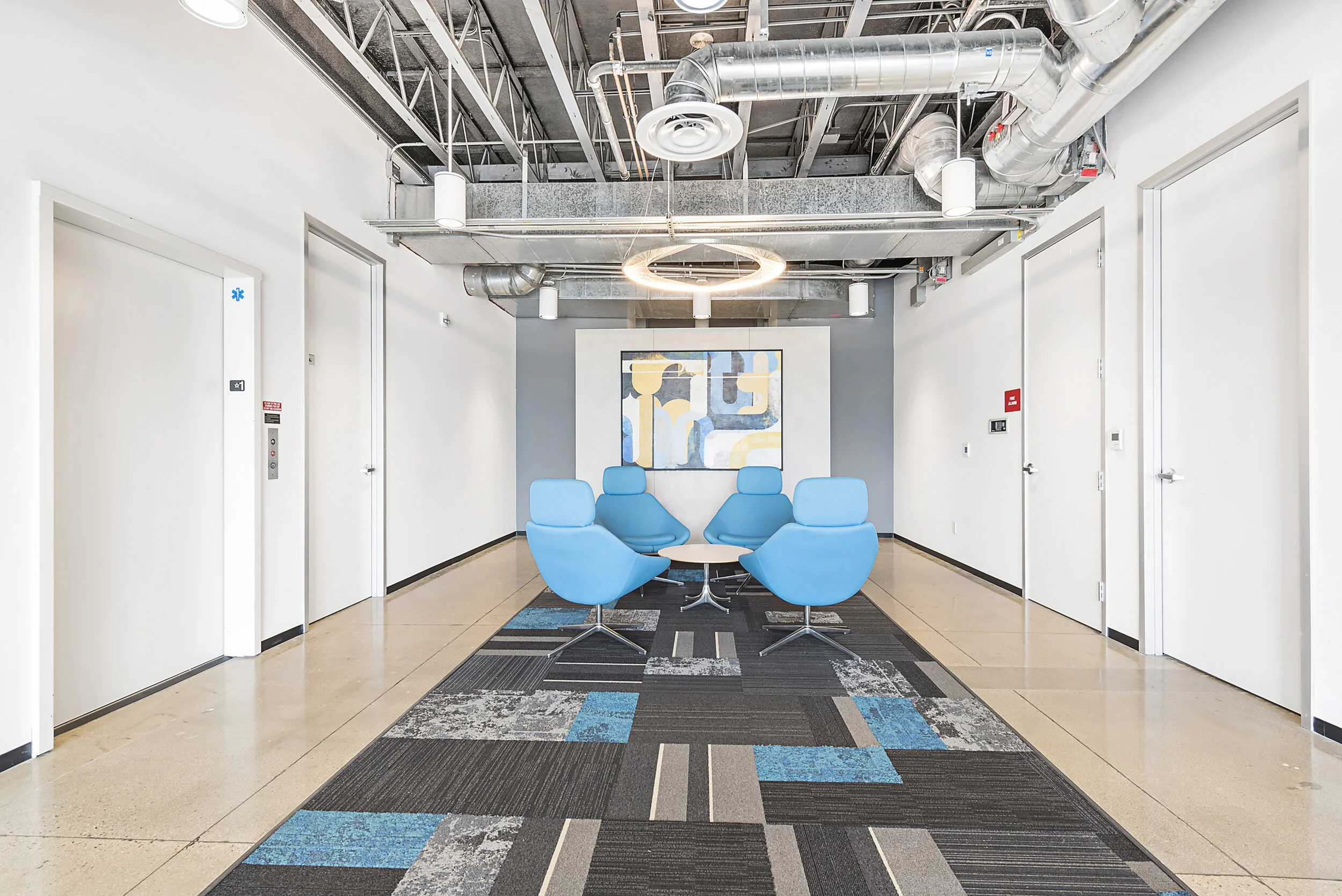 Waiting area with four blue chairs and a round table, patterned carpet, white walls, abstract artwork, and exposed ceiling ducts.