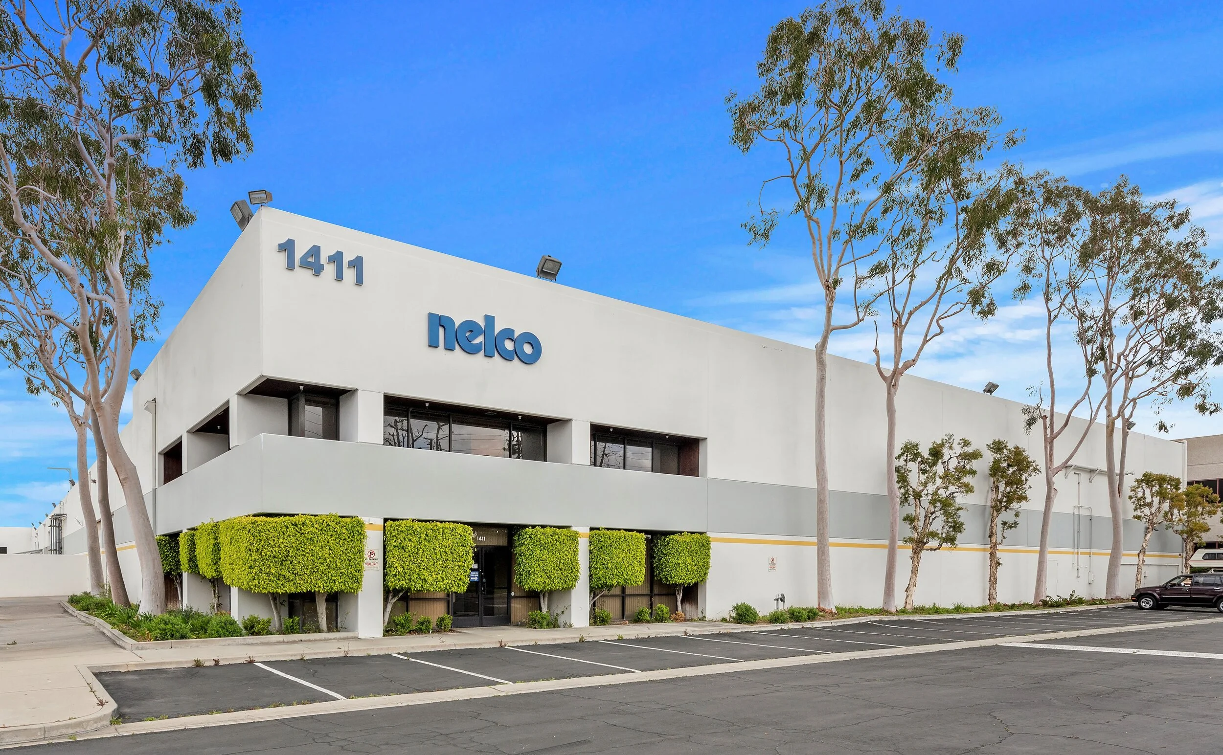 Large white building with blue 'nelco' sign, surrounded by trees and a parking lot, under a blue sky.