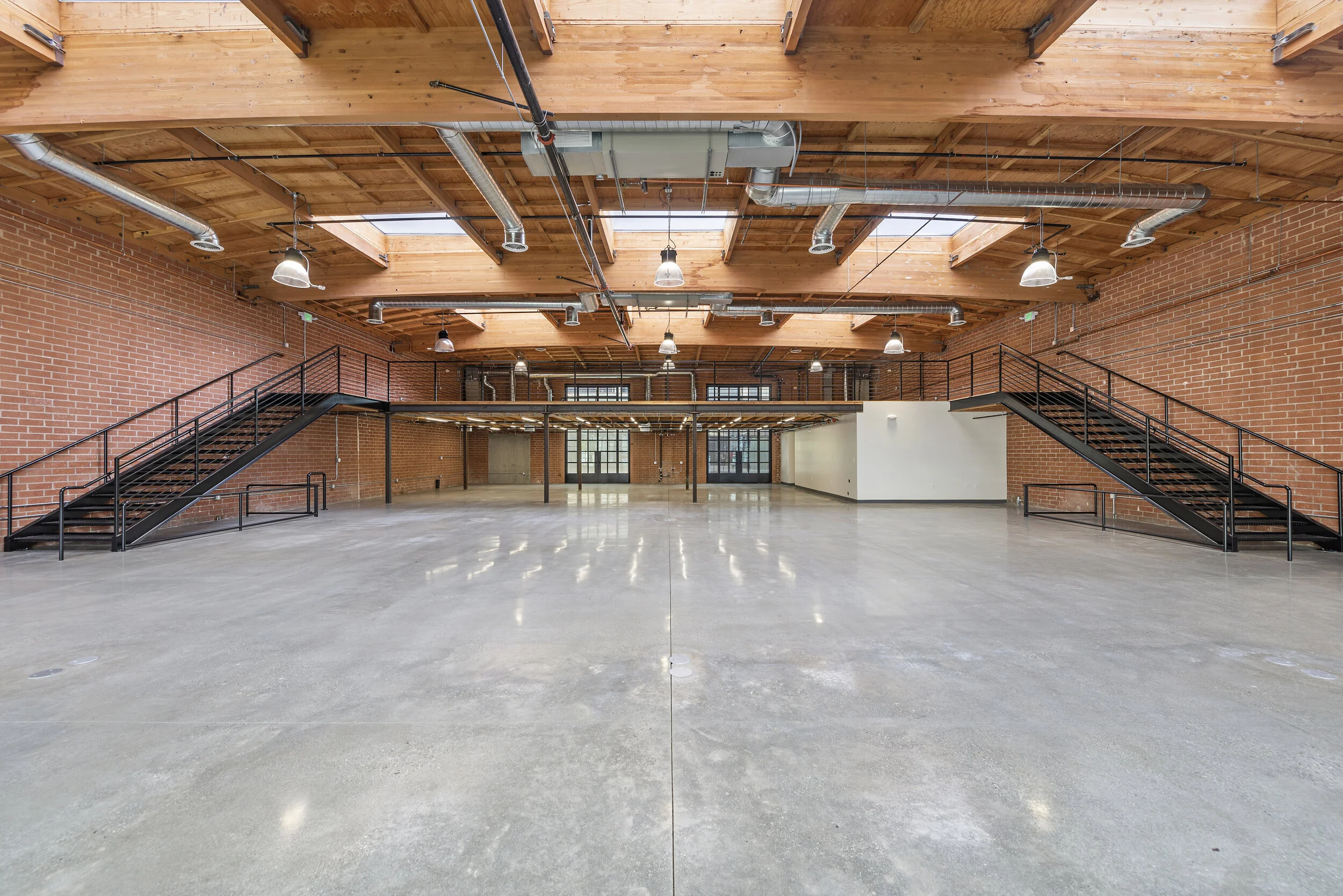 Empty industrial-style commercial space with polished concrete floors, exposed brick walls, wooden ceiling with ductwork, skylights, black metal staircases leading to a mezzanine, and large windows.