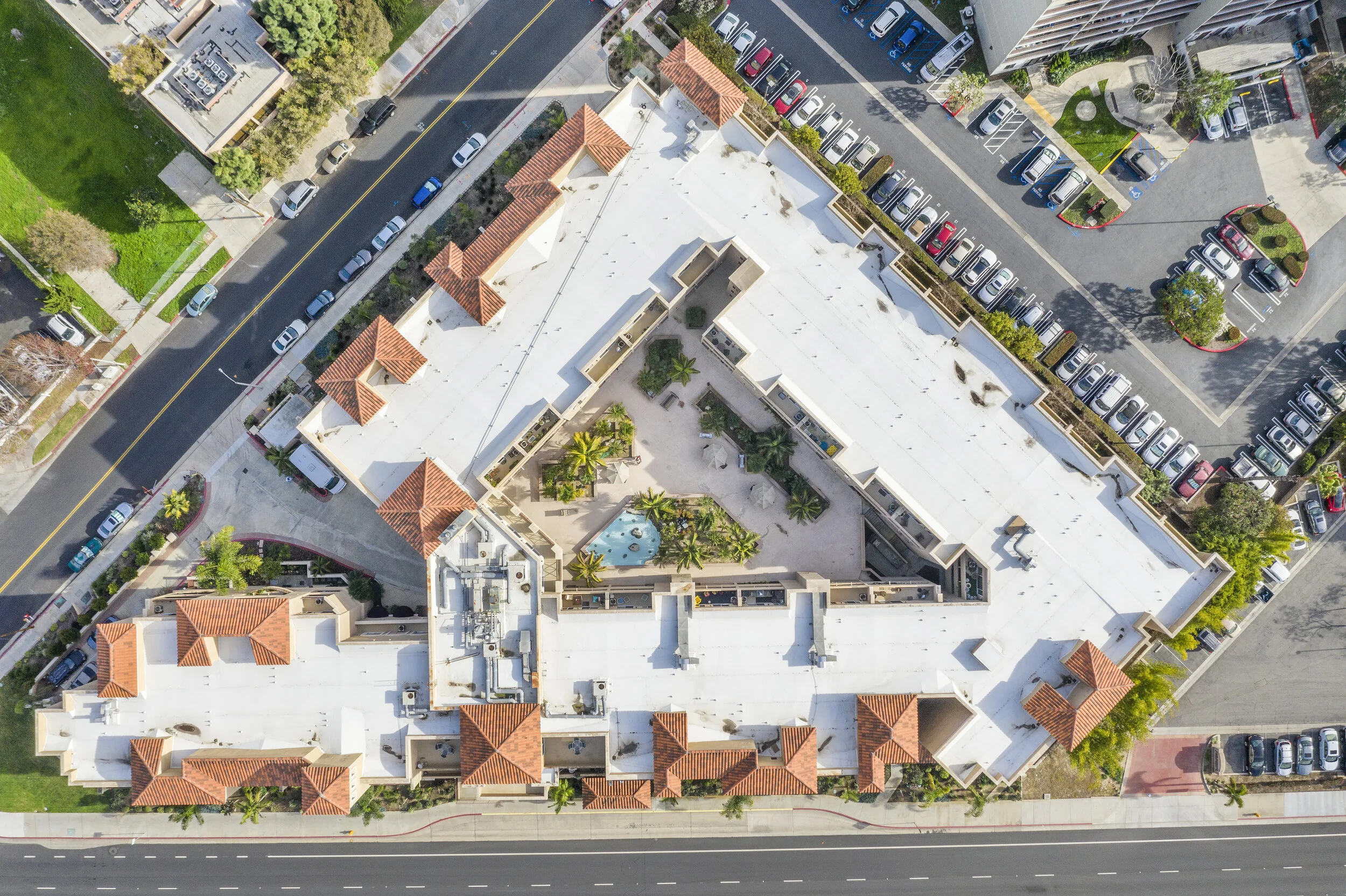 An aerial view of a large multi-story building with a rooftop pool and lounge area, surrounded by parking lots, roads, and landscaped greenery.