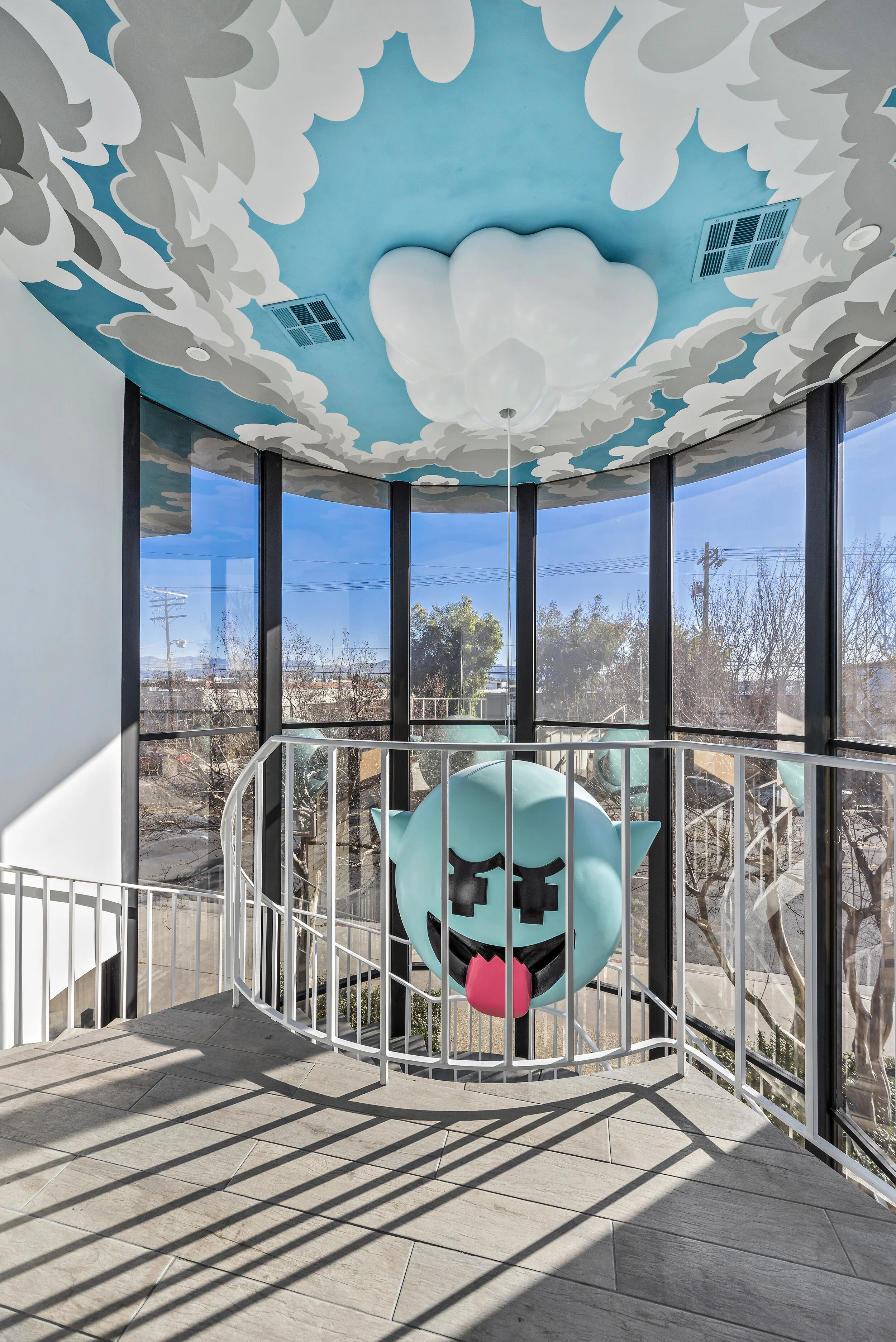 Interior view of a glass-enclosed space with a ceiling painted with clouds and a large cloud-shaped light fixture. There is a large, cartoonish, blue, smiling creature with black square eyes and a pink tongue hanging out, positioned behind a white ra