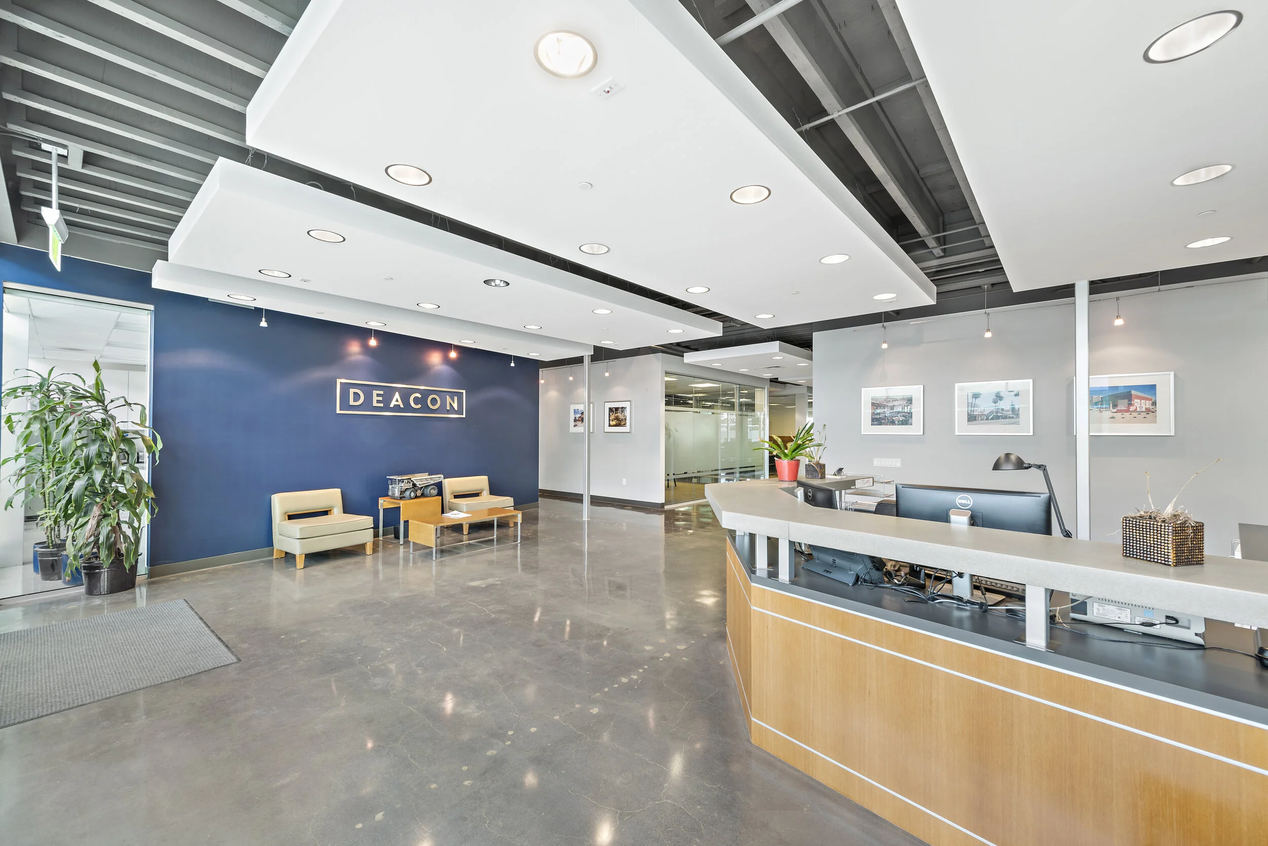 Modern office reception area with a blue accent wall displaying the company name 'Deacon', a service counter with computer monitors, potted plants, framed artwork on white walls, and seating with two beige chairs and a small table.
