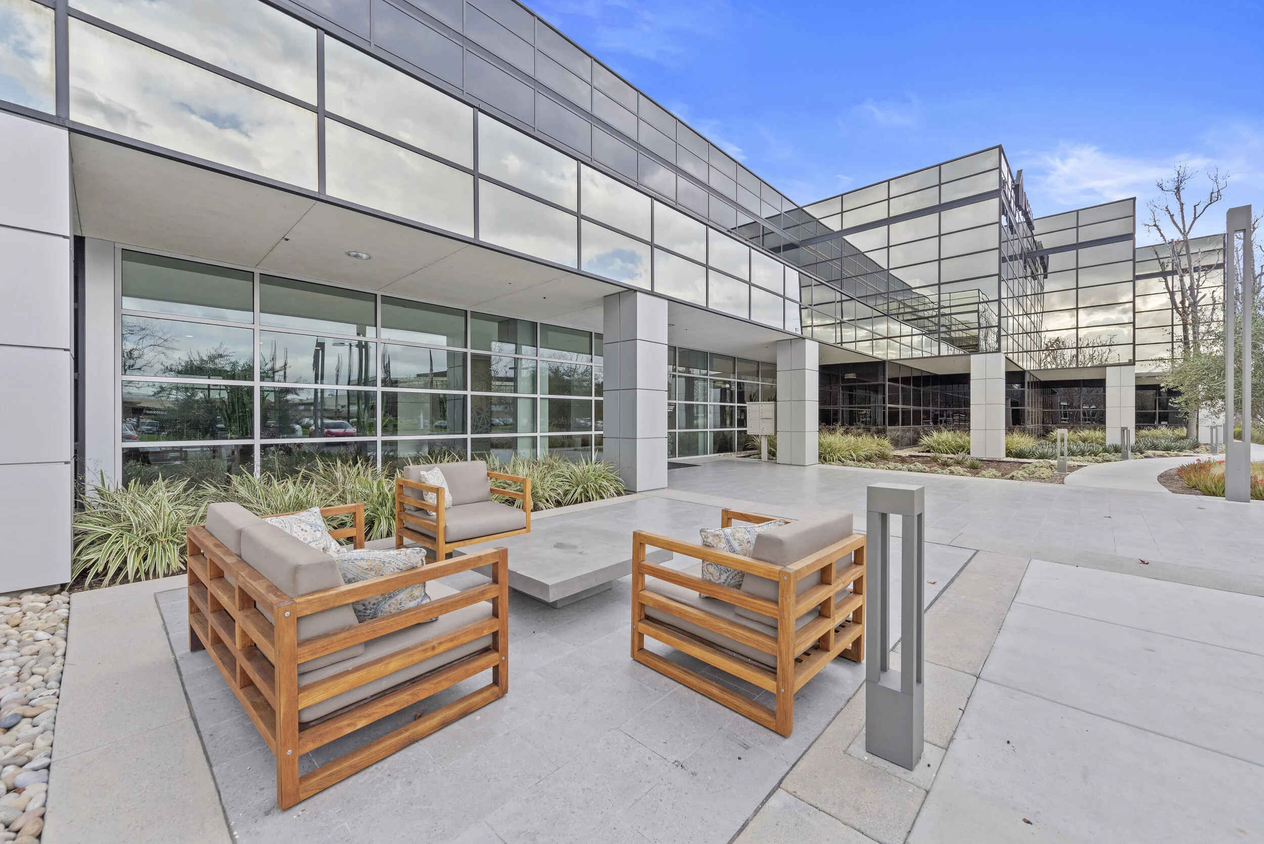 Modern office building with large glass windows, outdoor seating area with wooden chairs and cushions, landscaped with plants and narrow pathways, under blue sky with some clouds.