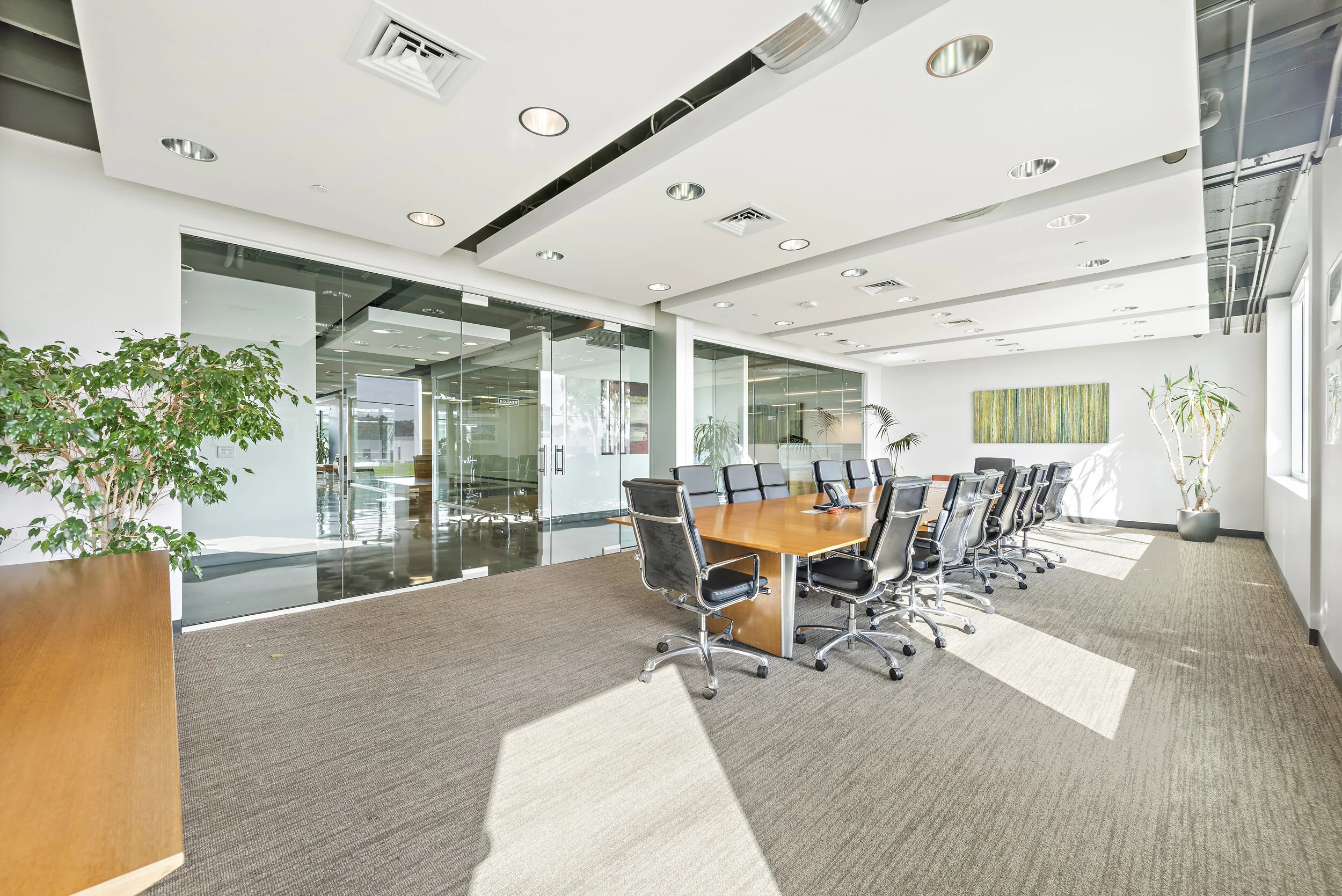 Empty modern conference room with a large wooden table, black office chairs, indoor plants, and glass walls.