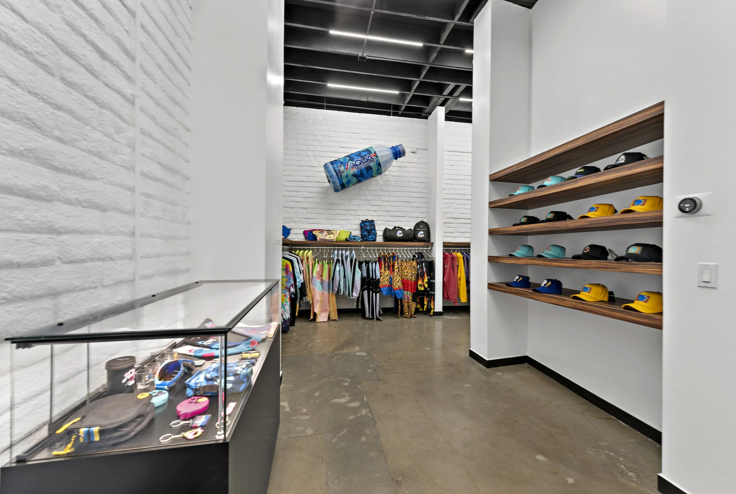Store interior with clothing on racks, shelves with caps, and a display case with accessories, white brick walls, wooden shelves, and a floating inflatable soda bottle decoration.