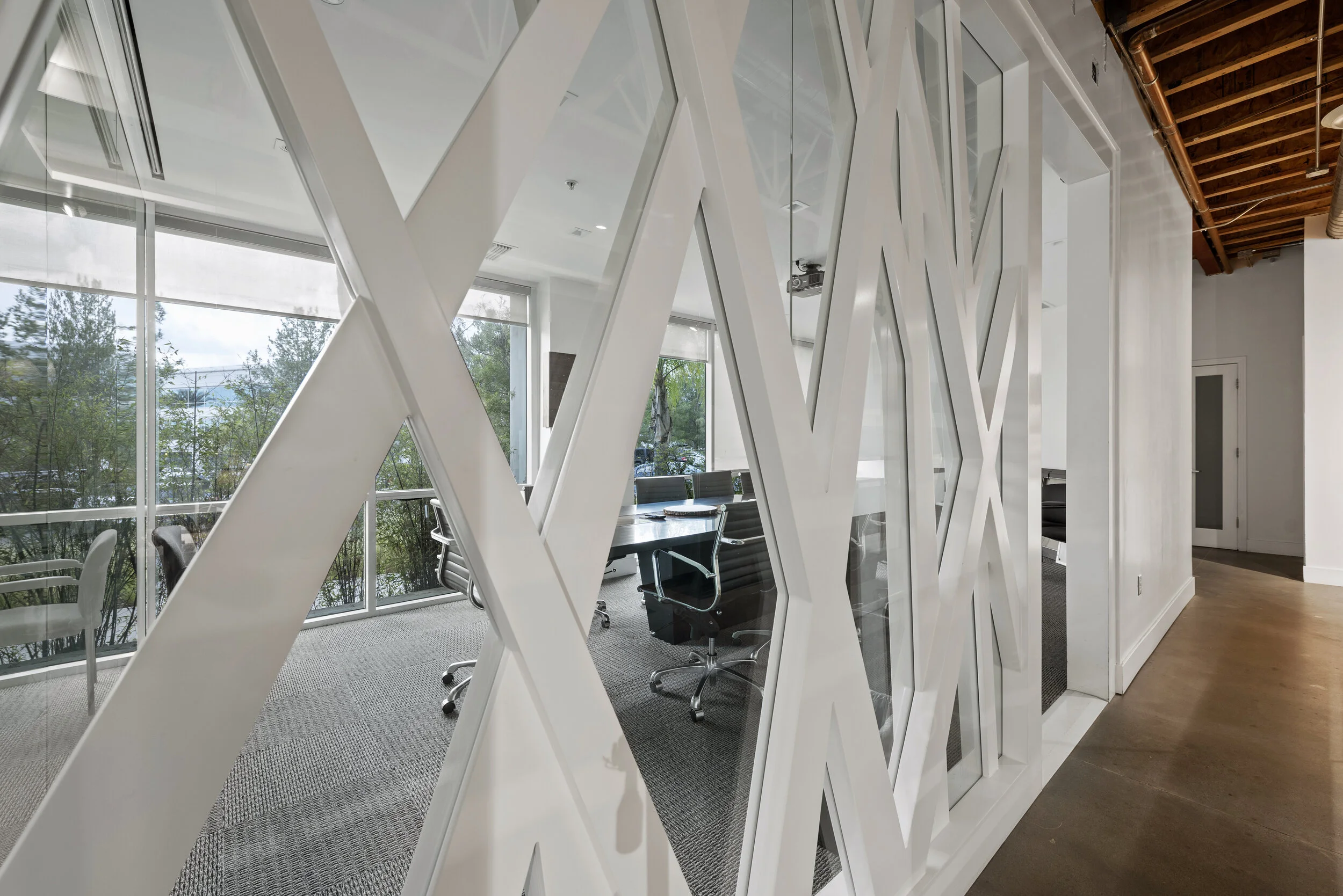 Modern office conference room with large glass windows, black chairs, and a wooden ceiling, viewed through a decorative white geometric partition.