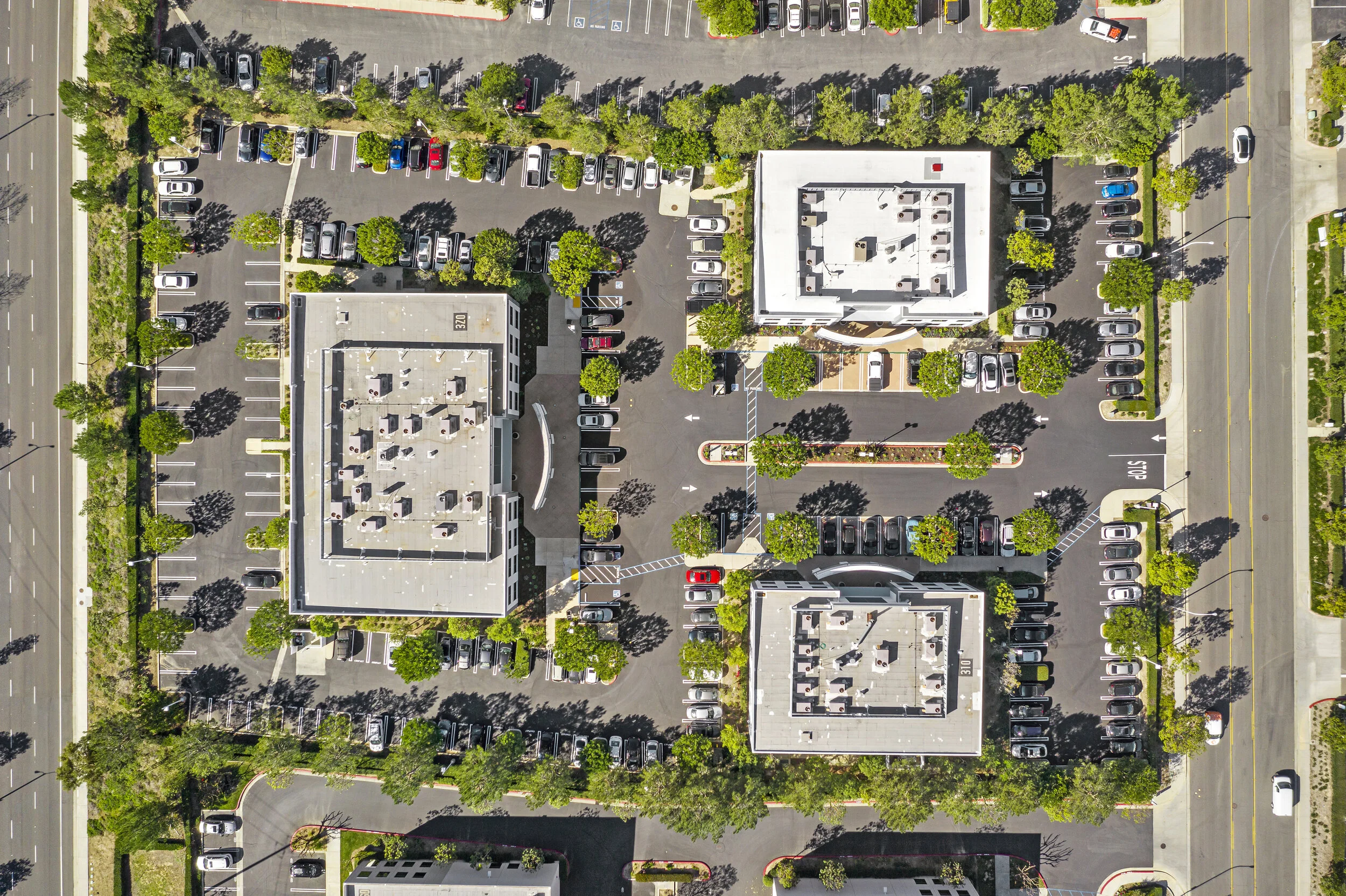 An aerial view of a parking lot with adjacent buildings, surrounded by trees and roads.