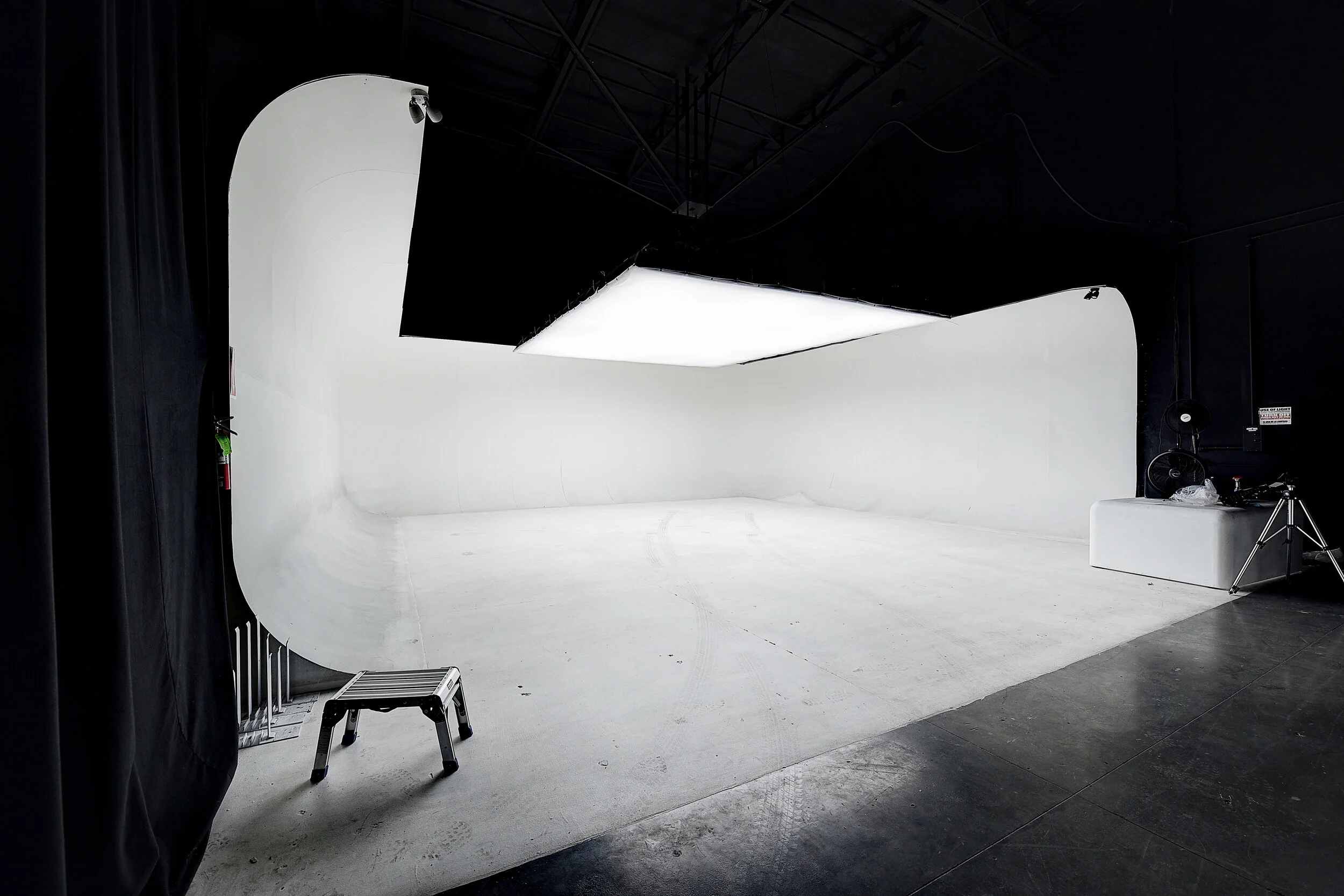 A photography studio with white cyclorama wall, large LED light panel overhead, and black side curtains.