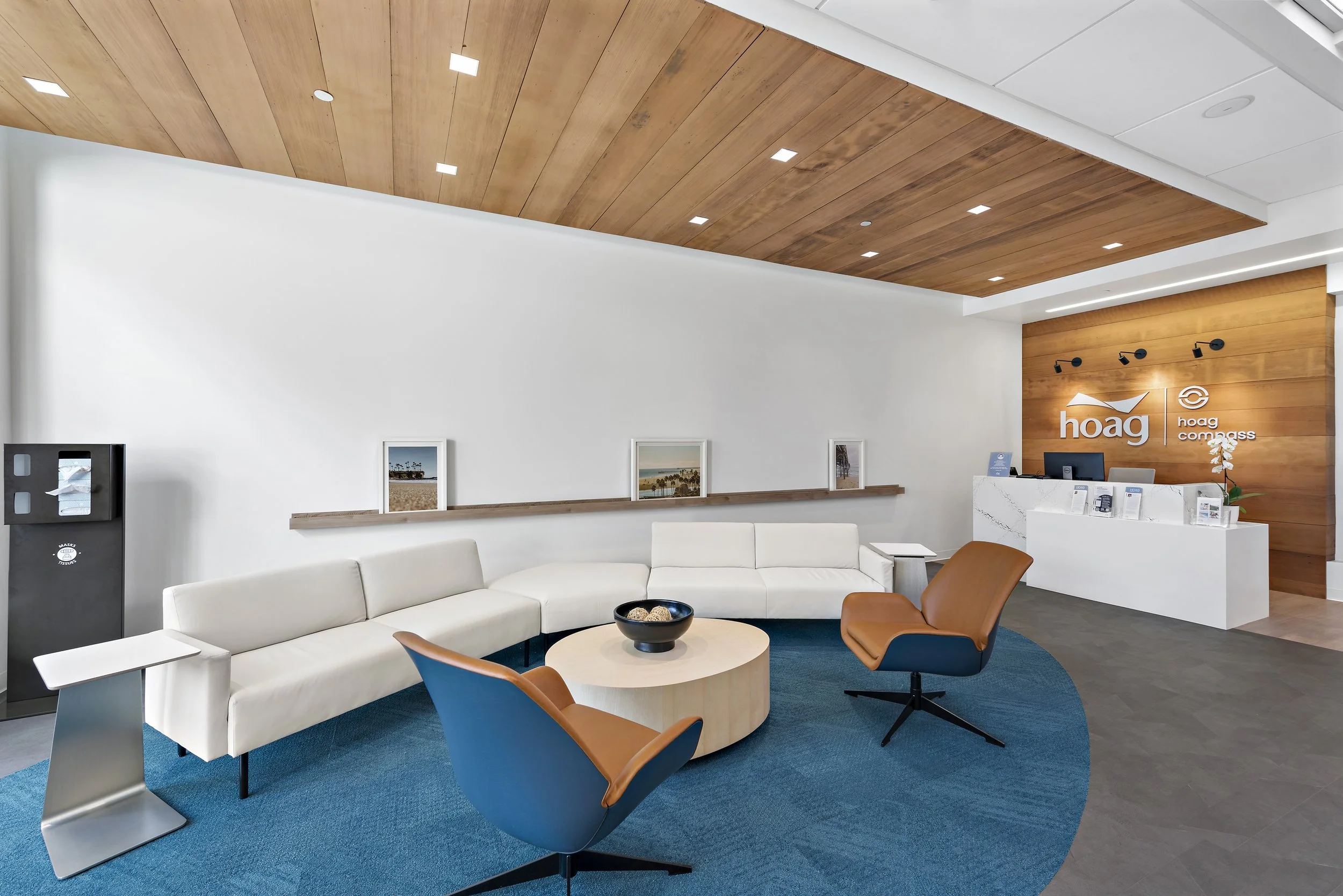 Modern reception area with white sectional sofa, two brown lounge chairs, a round coffee table, and images on a picture ledge. Branding for 'hoag' and 'hoag compass' on a wooden accent wall.